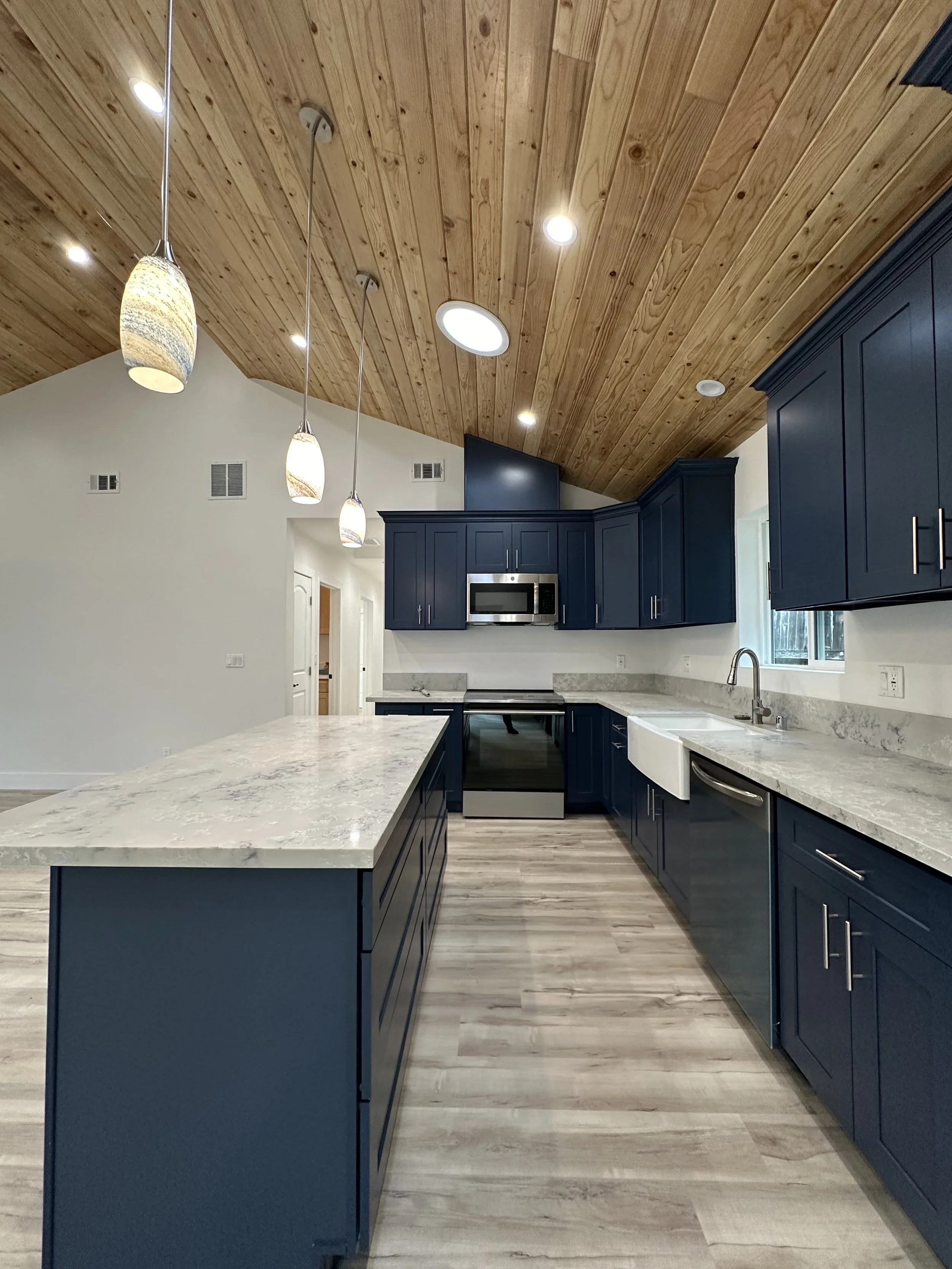 Modern kitchen with blue cabinets, a marble countertop island, stainless steel appliances, wooden ceiling with pendant lights, and a large window.