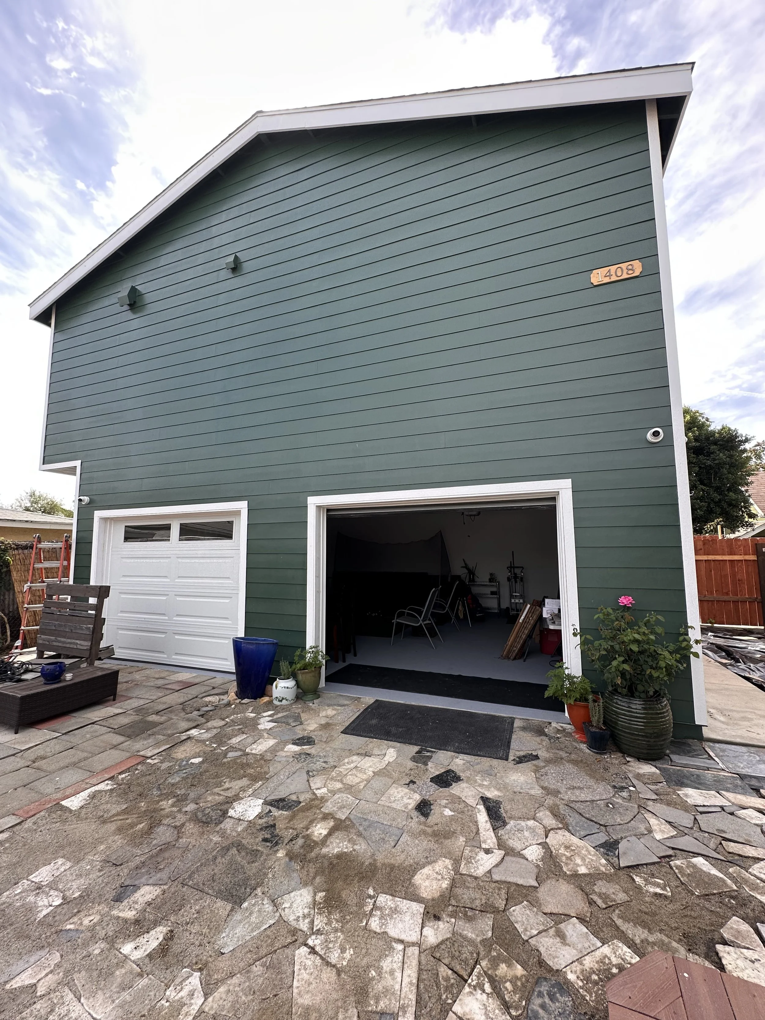 The back of a green two-story garage with one open door and one closed door, a brick stone patio, potted plants, and outdoor furniture.