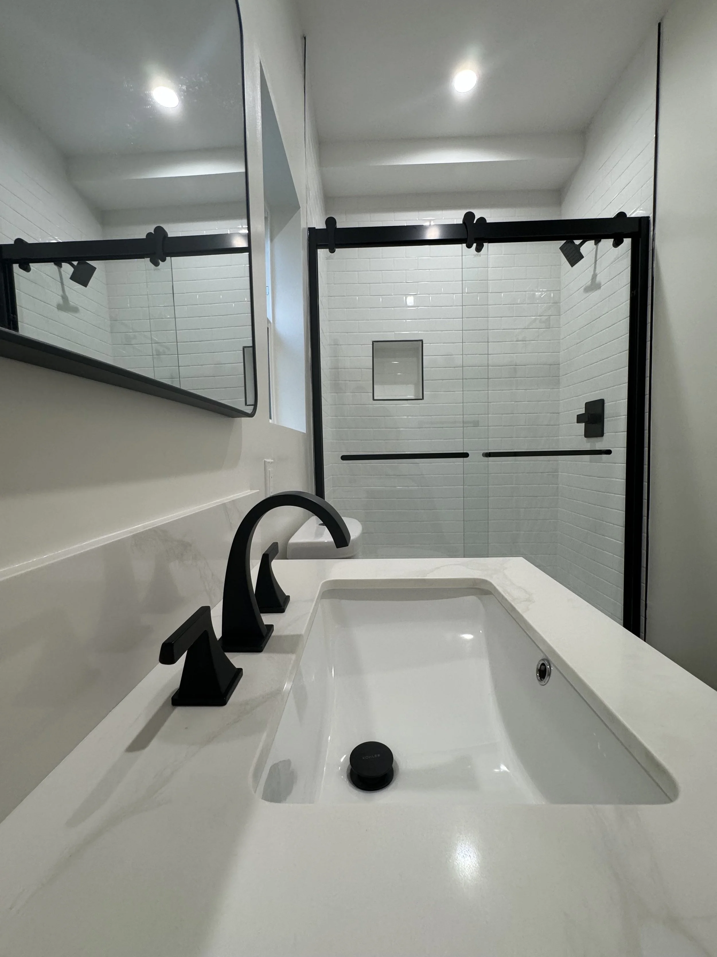Modern bathroom with white vanity, black faucets, and a glass-enclosed shower with white tile walls.