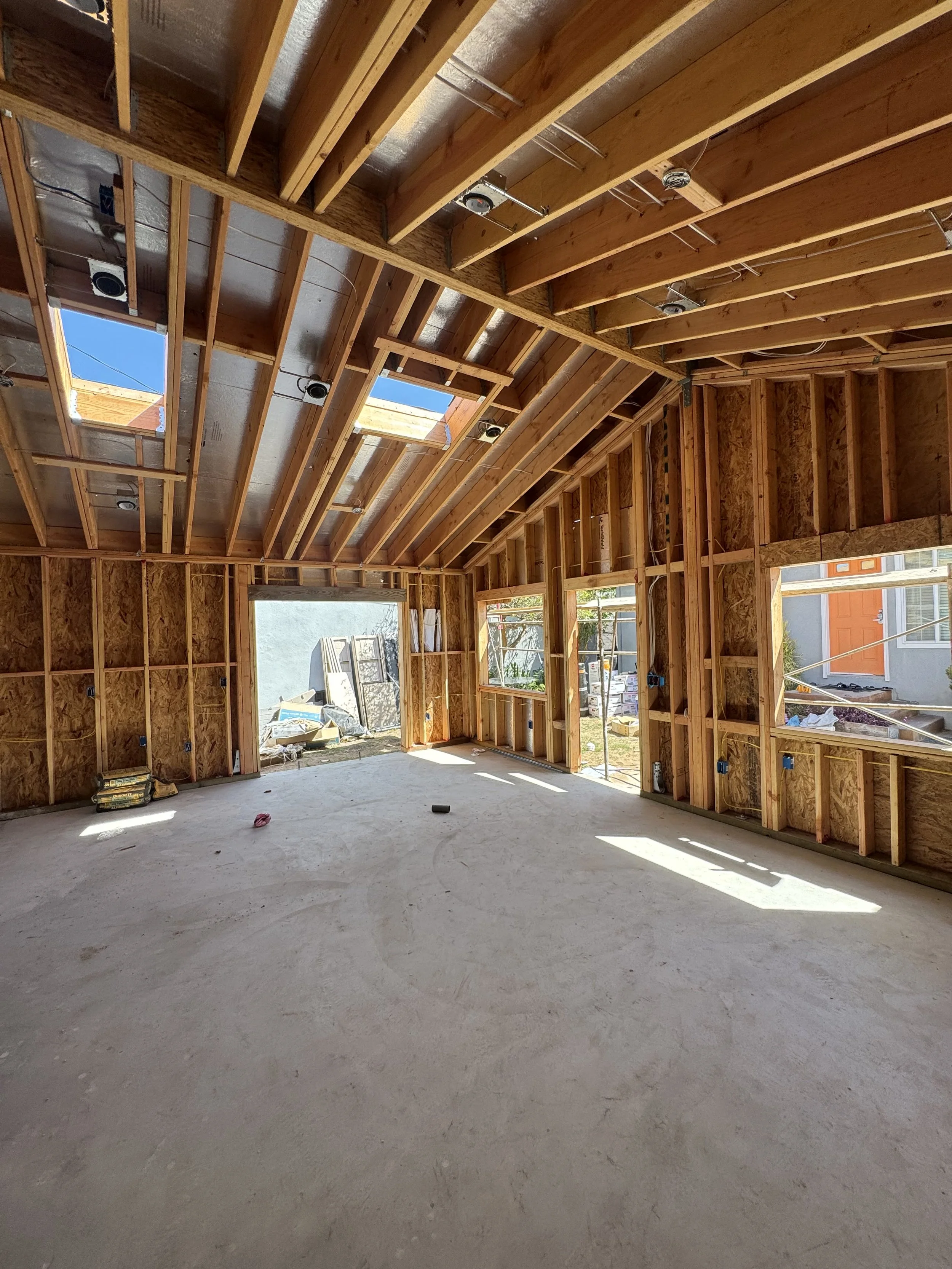Interior of a house under construction with exposed wooden framing, roof trusses, and skylights. The floor is unfinished concrete, and windows are installed. Outside, construction materials and tools are visible.