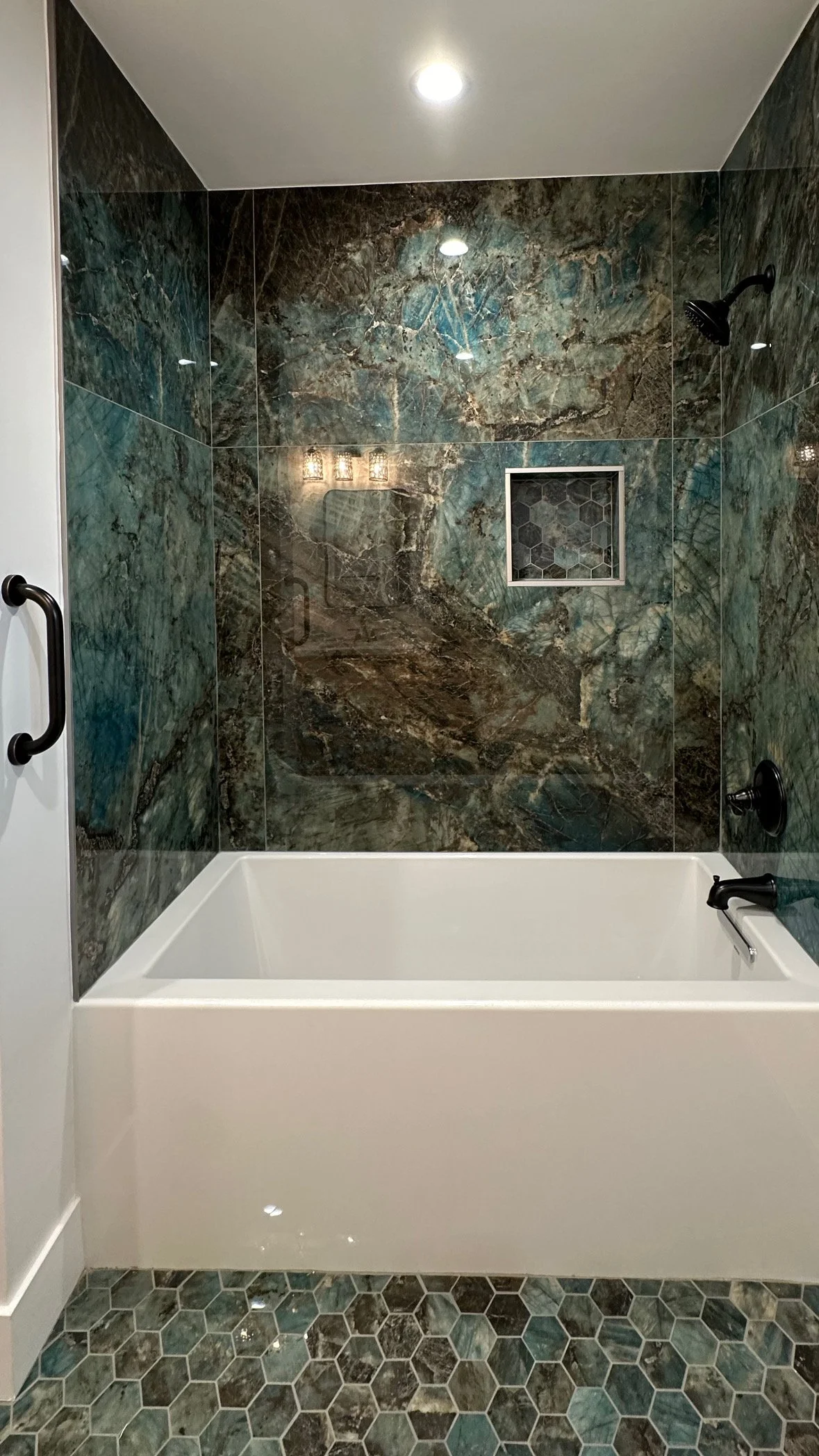 Bathroom shower with dark green and brown marble tiles, a small square niche, black fixtures, and a white bathtub.