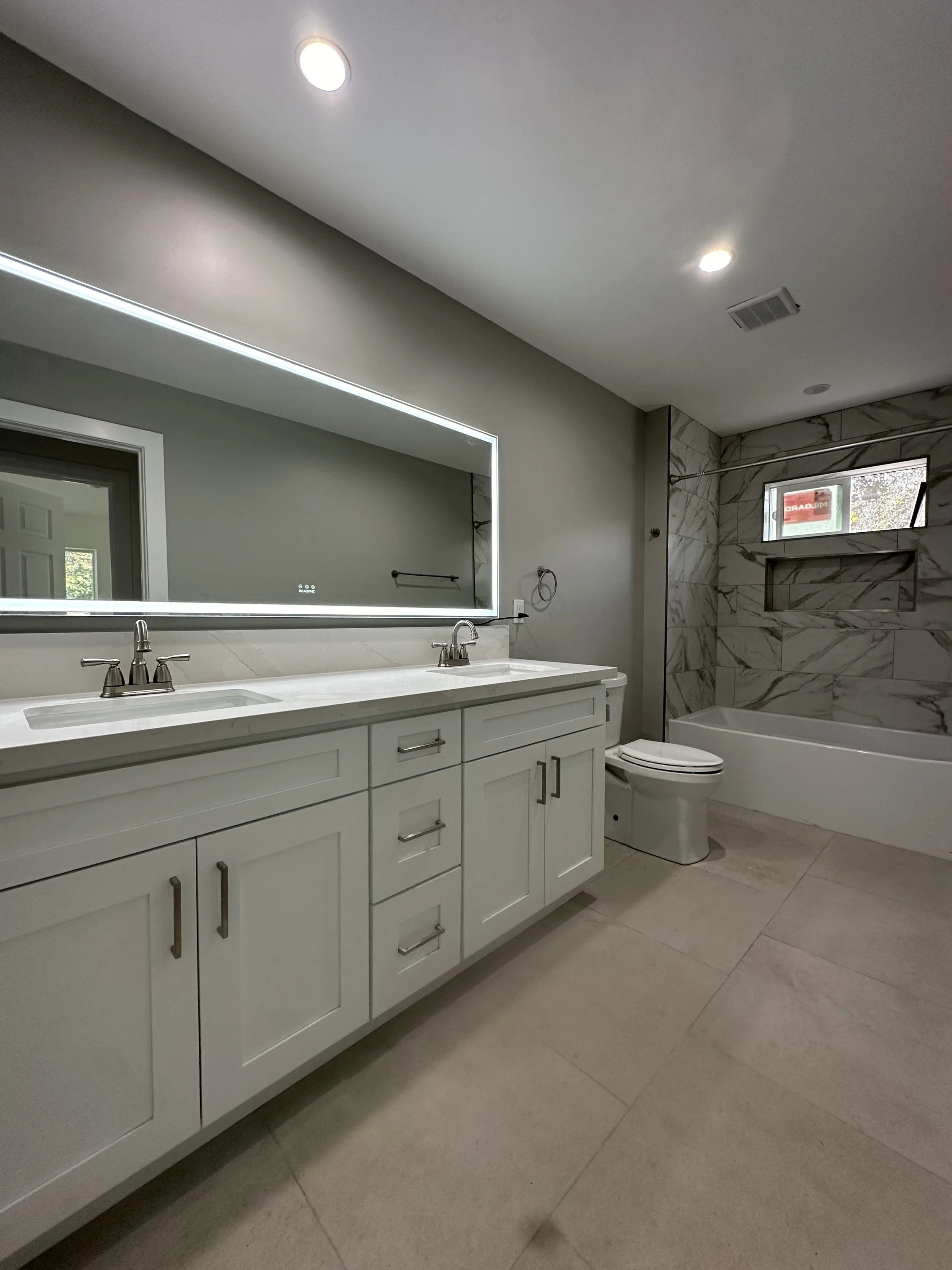 Modern bathroom with double vanity, large illuminated mirror, bathtub with marble wall, toilet, beige tile floor, ceiling lights, and window.