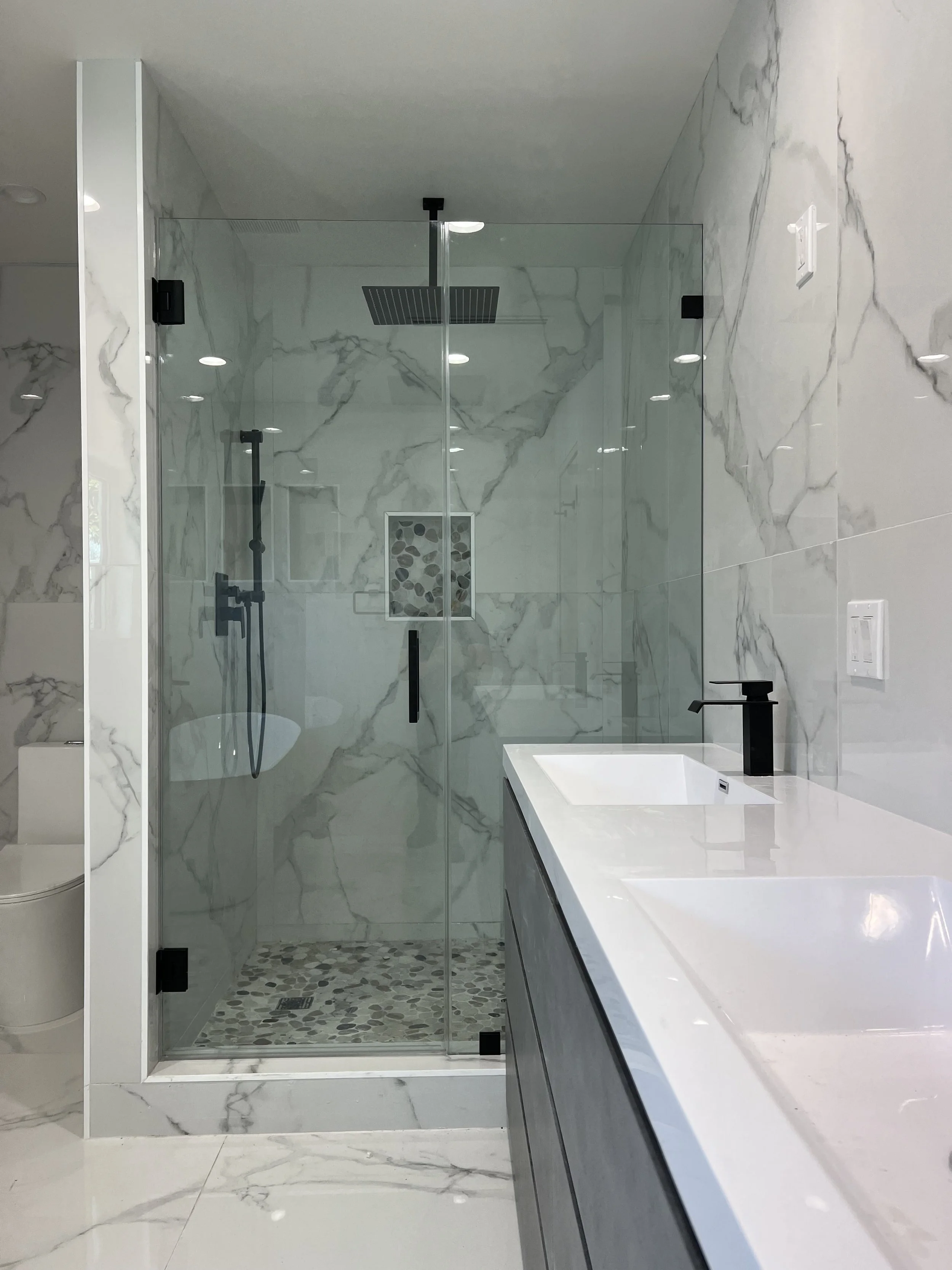 Modern bathroom with marble walls and flooring, glass shower enclosure with pebble tile floor, black fixtures, and a white vanity with a black faucet.