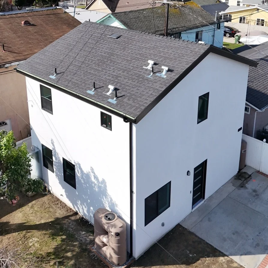 A modern two-story white house with a gray shingle roof, several windows, and a driveway in the front. A fenced backyard with a tree and outdoor storage tank is visible.