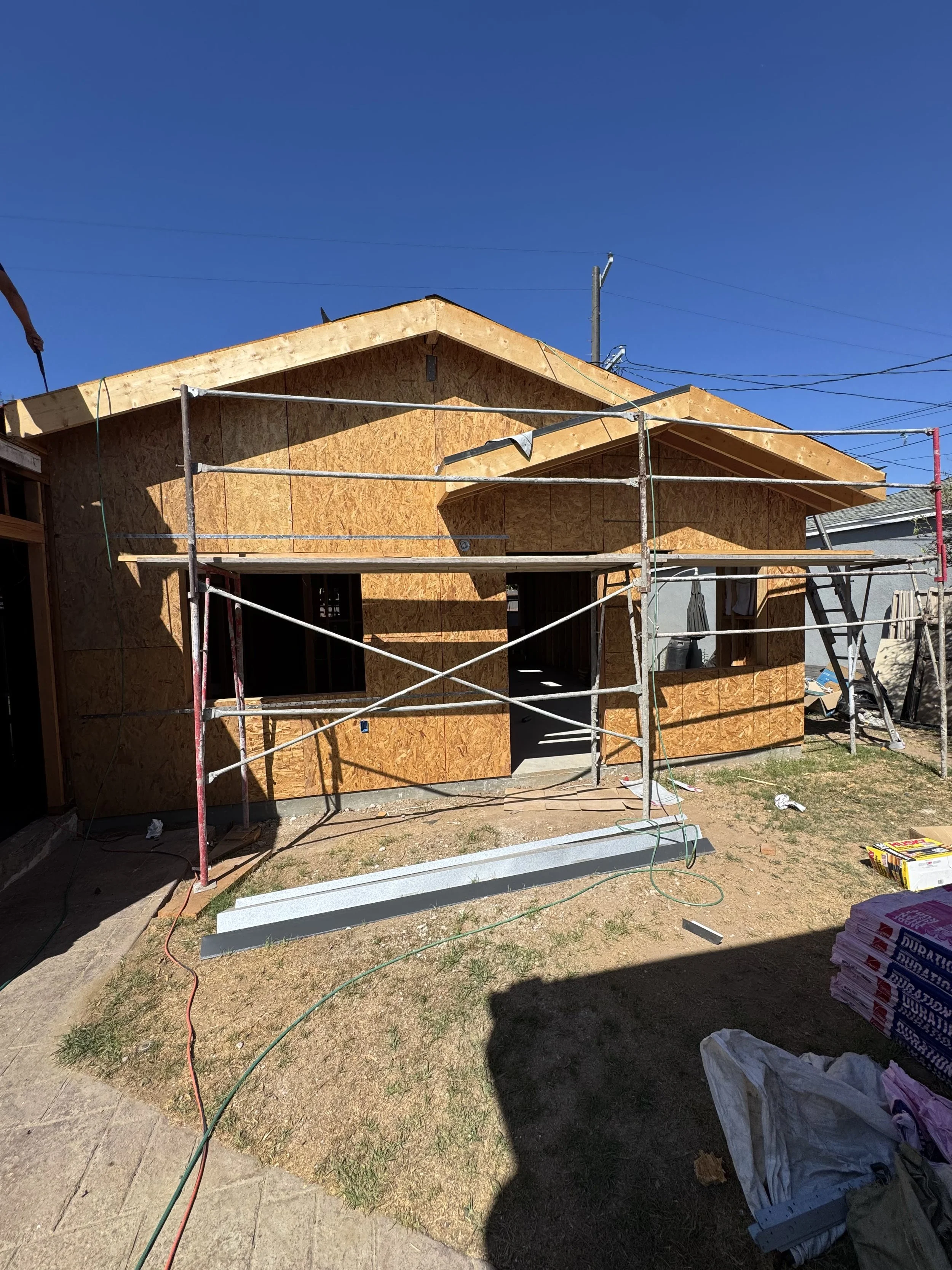House under construction with exterior walls covered in plywood, scaffolding around it, and construction materials on the ground under a clear blue sky.