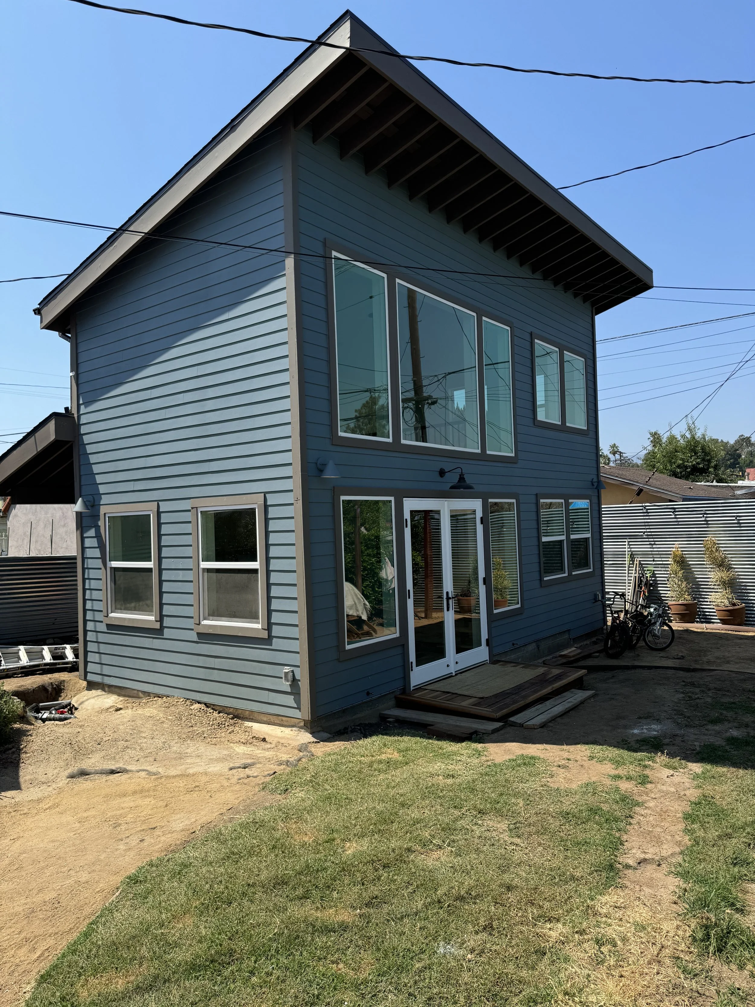 Newly constructed blue multi-story house with large windows and a sliding glass door, surrounded by a yard with patchy grass and dirt, bicycles parked outside, and a backyard fence.