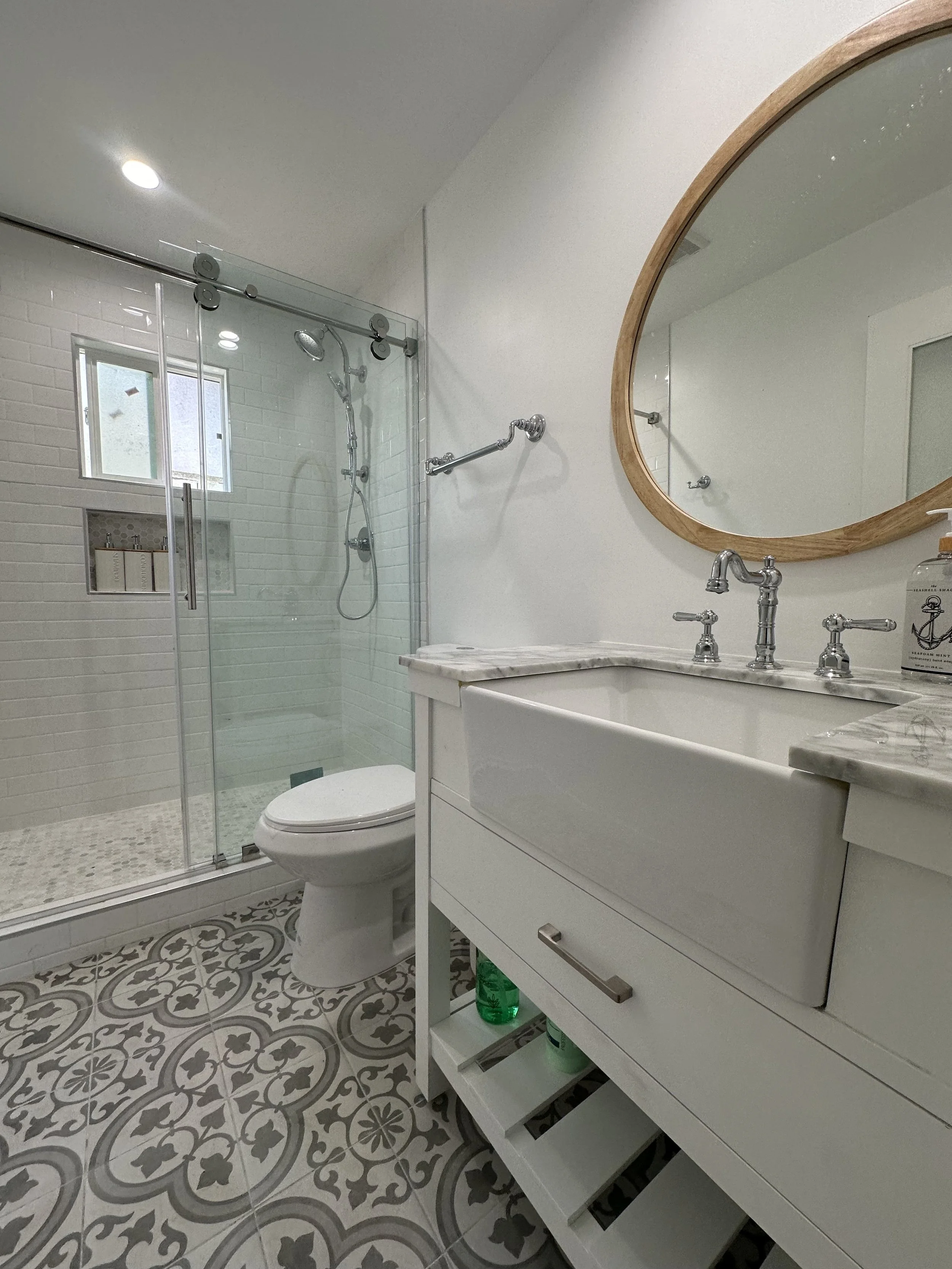 Modern bathroom with patterned floor tiles, white vanity with marble countertop, oval mirror with wooden frame, glass-enclosed shower with white subway tile, and small window for natural light.
