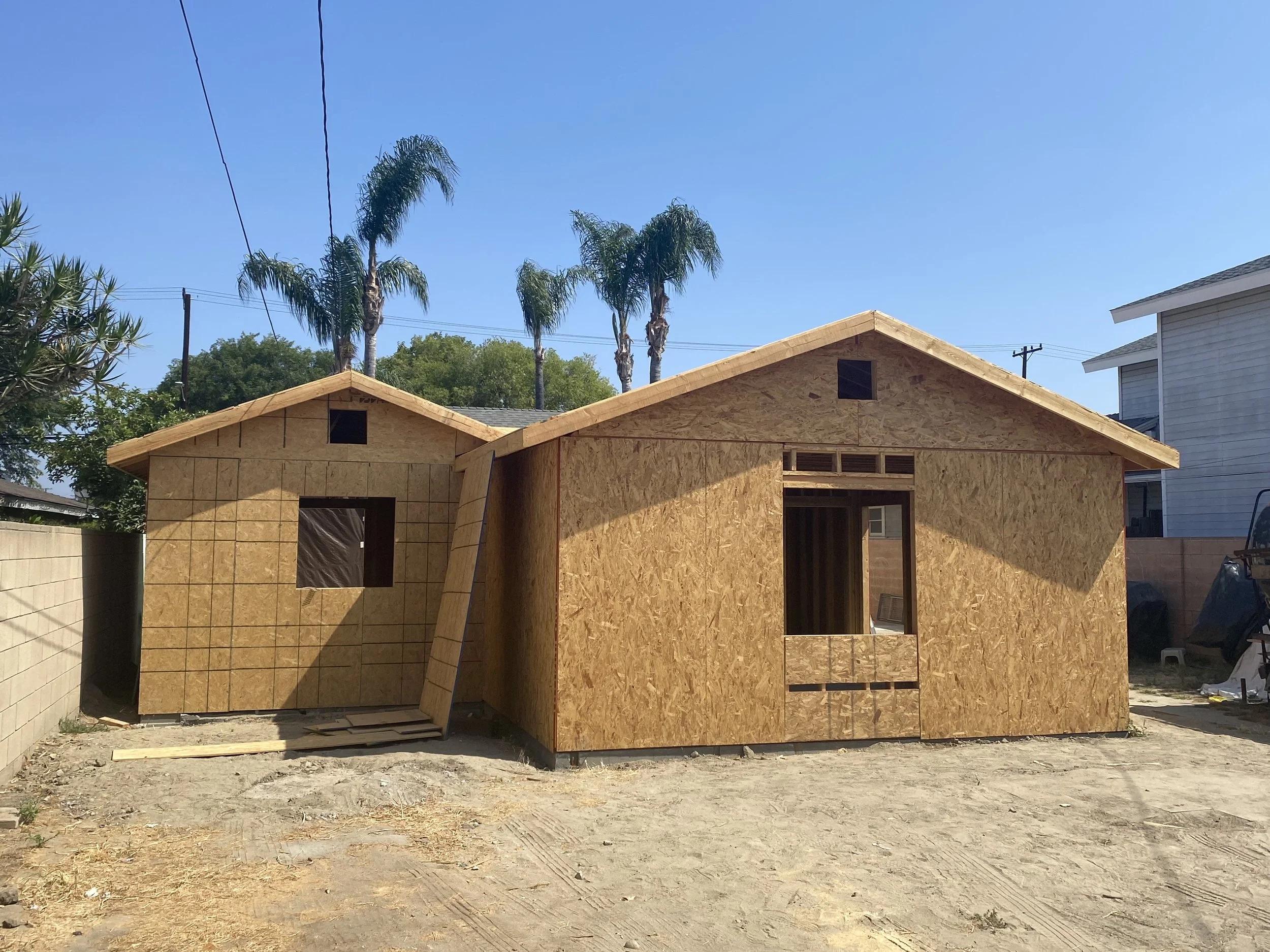 Two small houses under construction with unfinished wooden walls and no windows, set on a dirt lot with a cement block wall on the side and palm trees and a blue sky in the background.