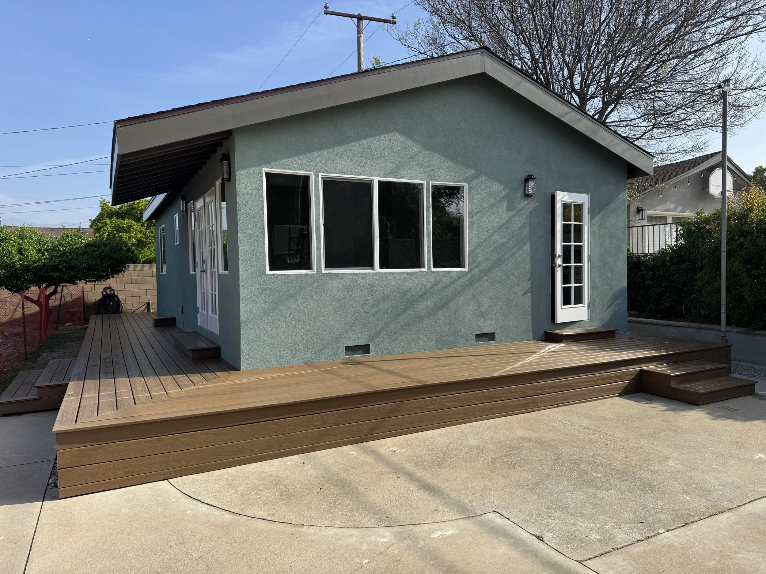 Backyard of a house with a wooden deck, light blue exterior walls, and white-framed windows and door, with steps leading up to the deck.