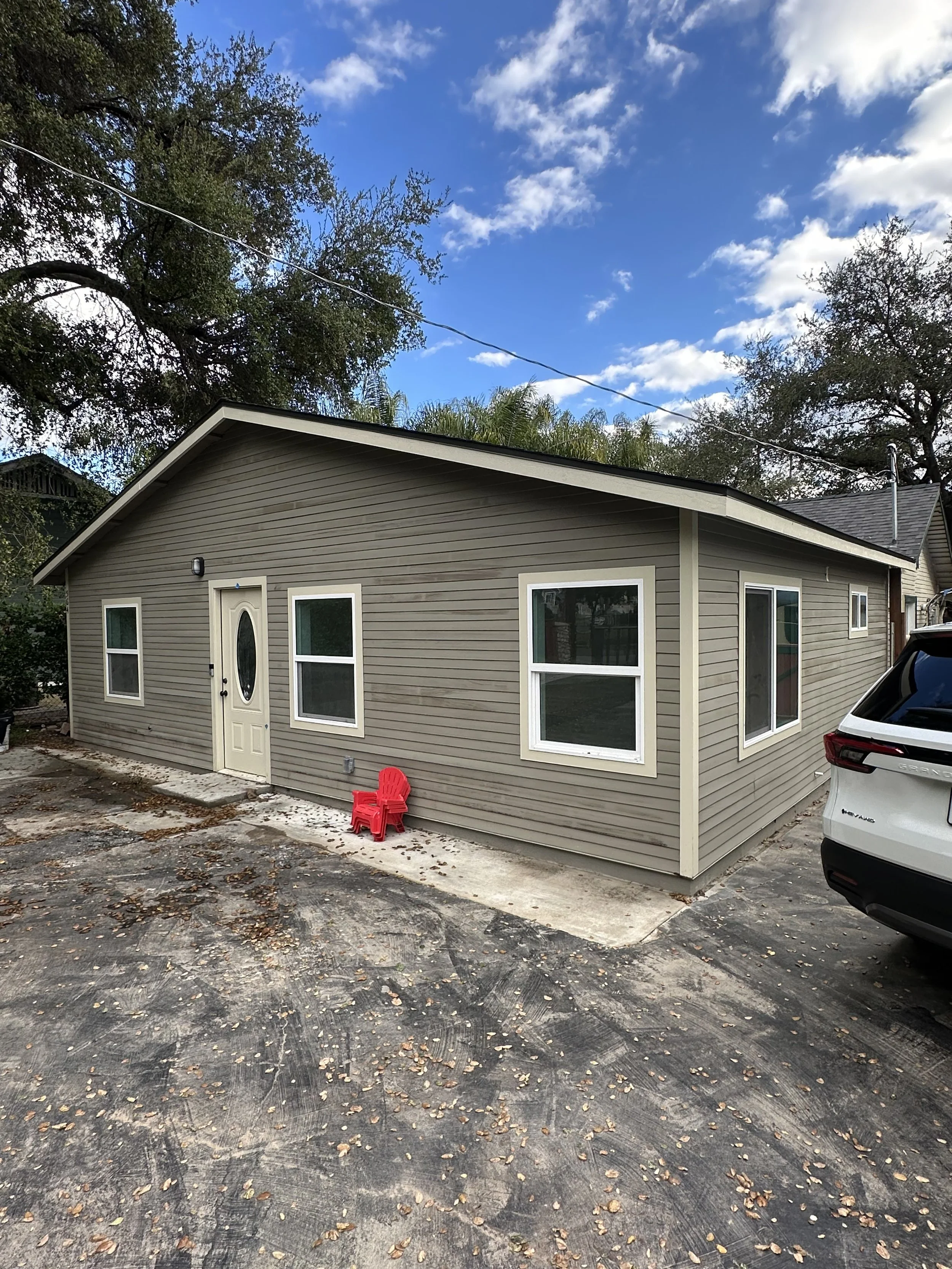 A small beige house with white trim, four windows, and a front door. There are two red chairs outside, on a concrete step. A white SUV is parked nearby. The ground is paved with asphalt and fallen leaves. Trees and a partly cloudy sky are in the back