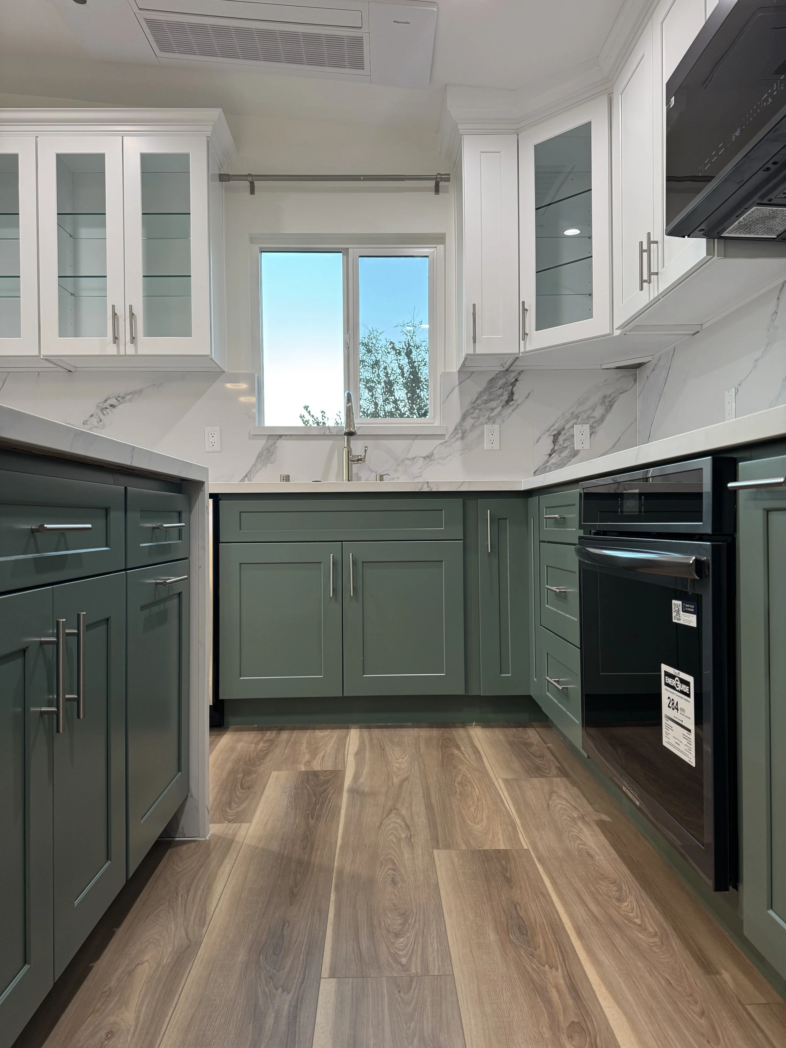 Modern kitchen with green lower cabinets, white upper cabinets, marble backsplash, wooden flooring, window above the sink, stainless steel fixtures, and a black oven.