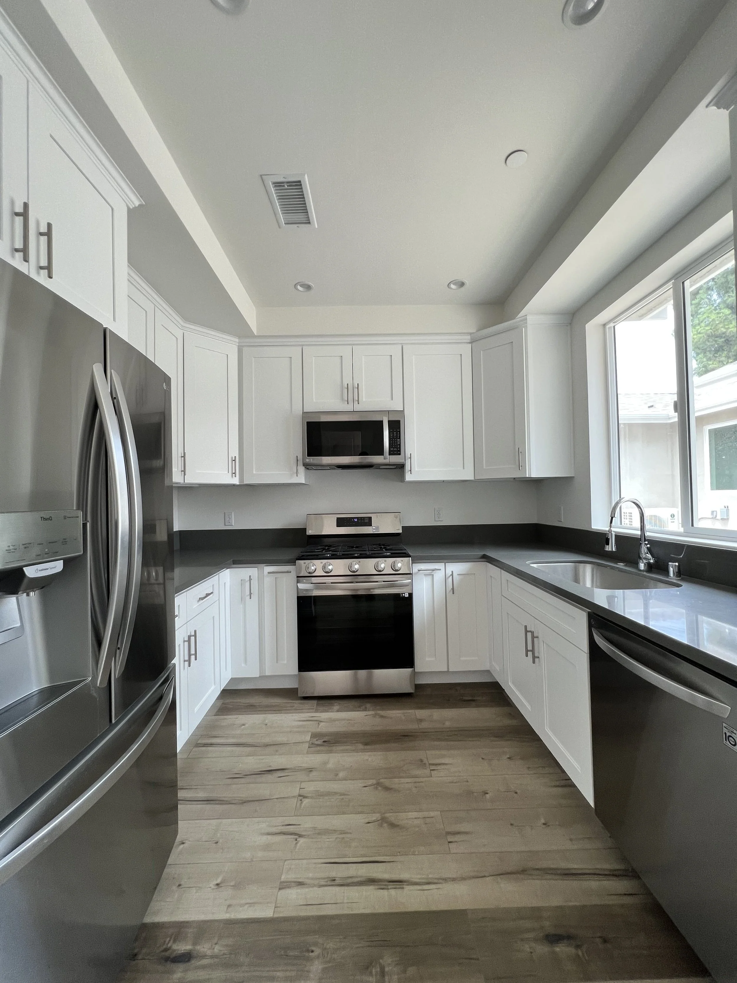 Modern kitchen with white cabinets, stainless steel appliances including a refrigerator, microwave, oven, and dishwasher. There is a large window above the sink, with light wood flooring.