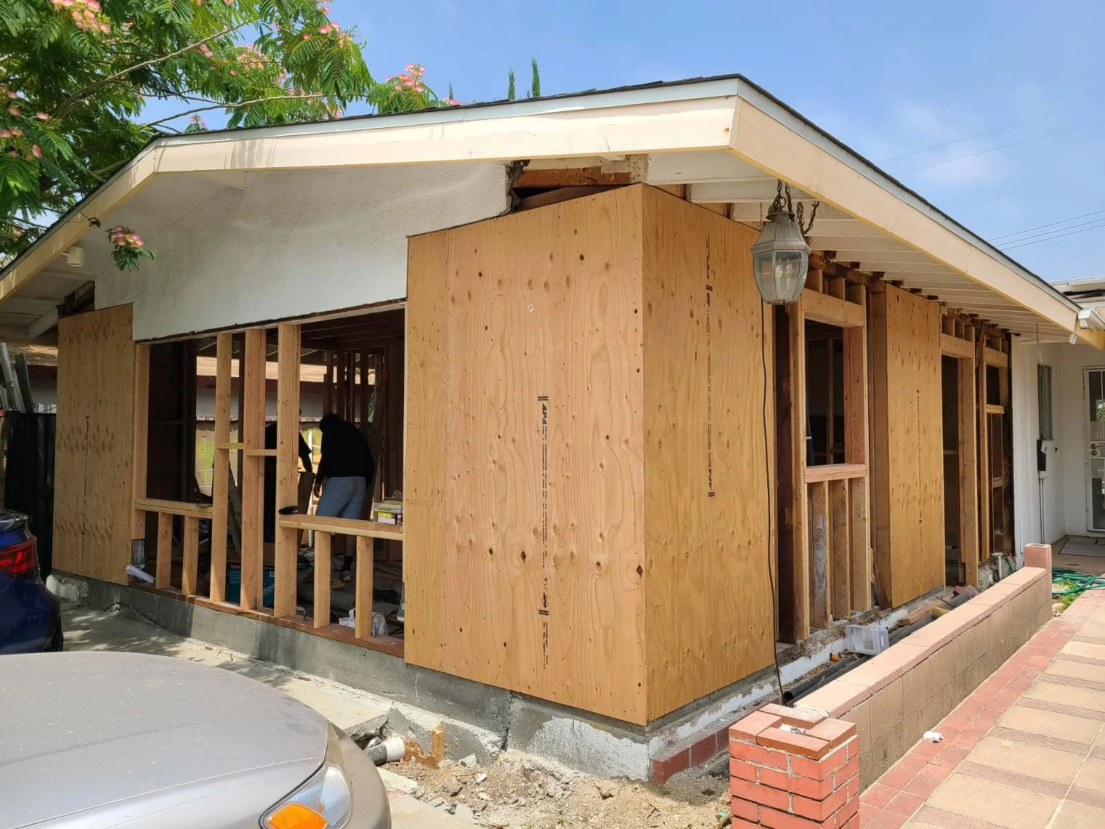 House under construction with plywood framing on the exterior walls, a white roof, and a person working inside. There is a car parked in front and a brick border along the sidewalk.