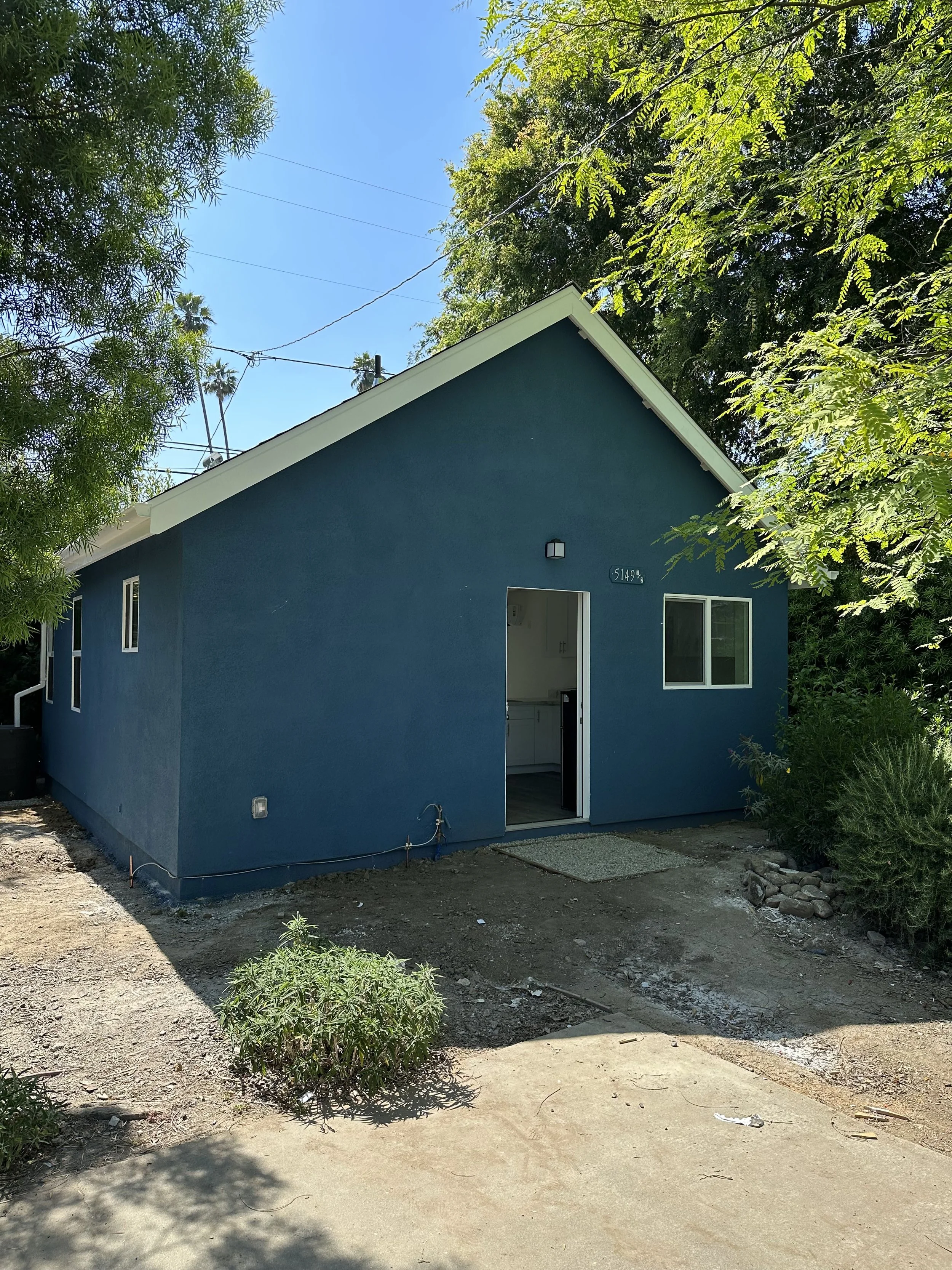 A small house painted dark blue with a white door open, showing a glimpse of the interior kitchen. The house has a sloped roof, two small windows on the side, and is surrounded by trees and bushes. The ground outside is dirt and concrete, with some r