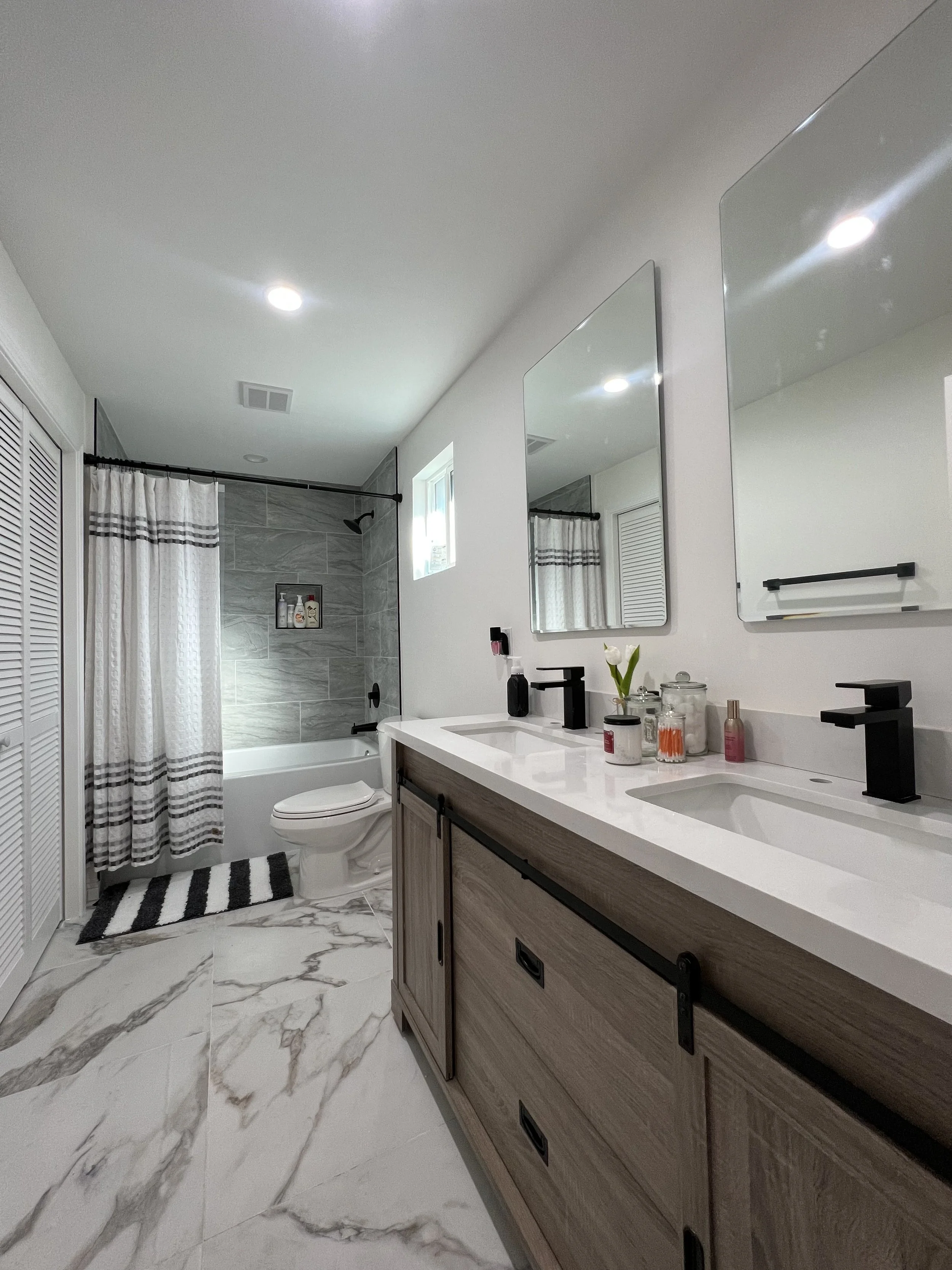 Modern bathroom with double vanity sink, large mirrors, and a walk-in shower with gray marble tiles. There are black fixtures, toiletries, a small window, and a striped rug in front of the toilet.