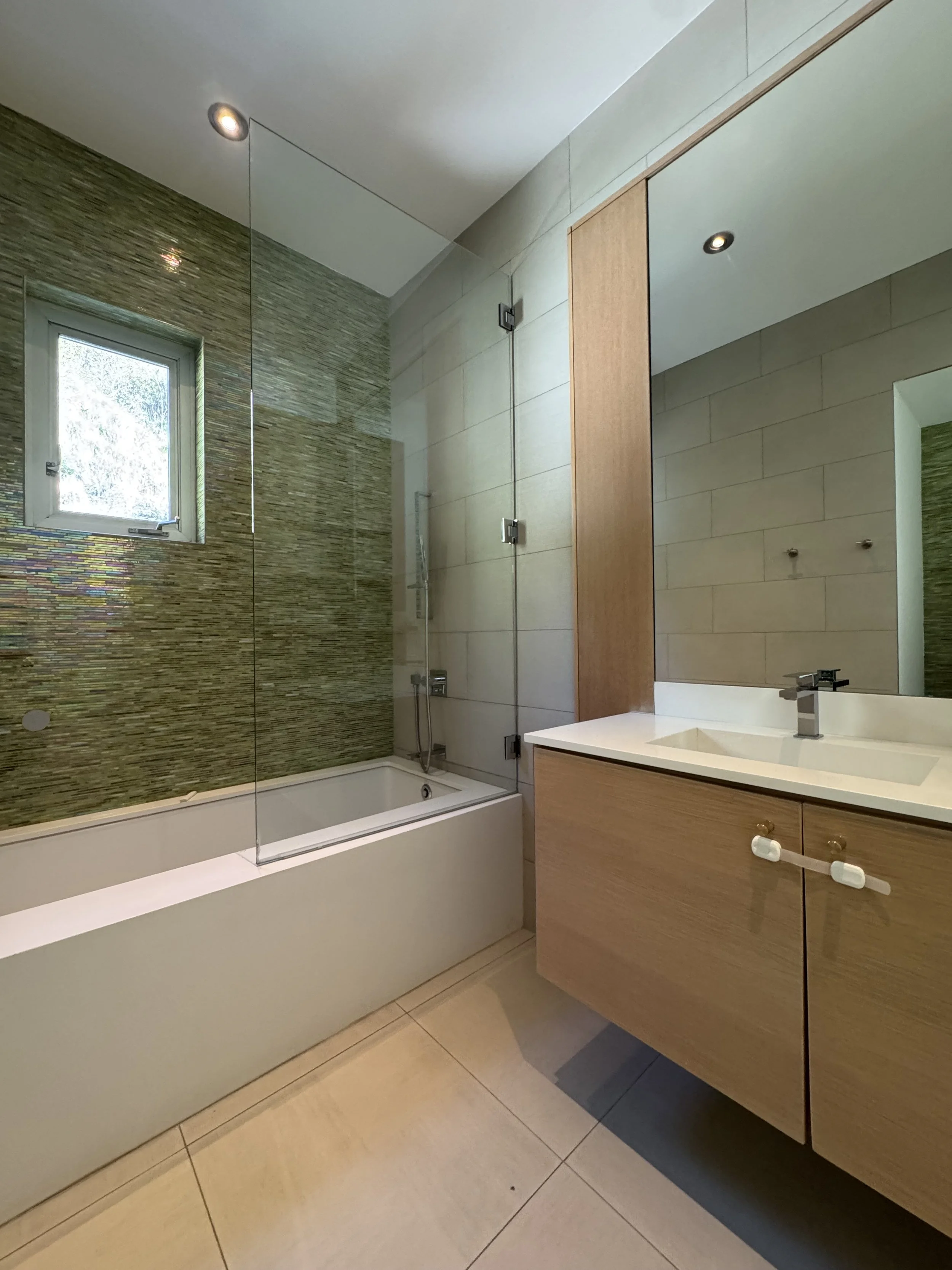 Modern bathroom with bathtub and glass shower partition, green tiled wall, small window, beige tiled floor, and wooden vanity with sink and mirror.