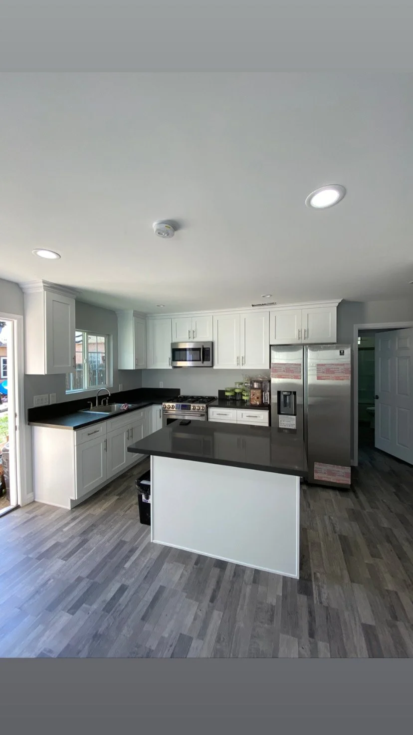 Modern kitchen with white cabinets, black countertops, stainless steel appliances including a refrigerator, microwave, and stove, with wooden flooring and a small kitchen island.