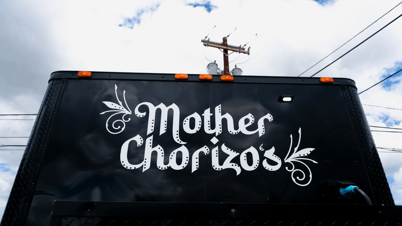 Black food truck with white decorative lettering that reads 'Mother Chorizos' and floral accents, under a partly cloudy sky with power lines overhead.
