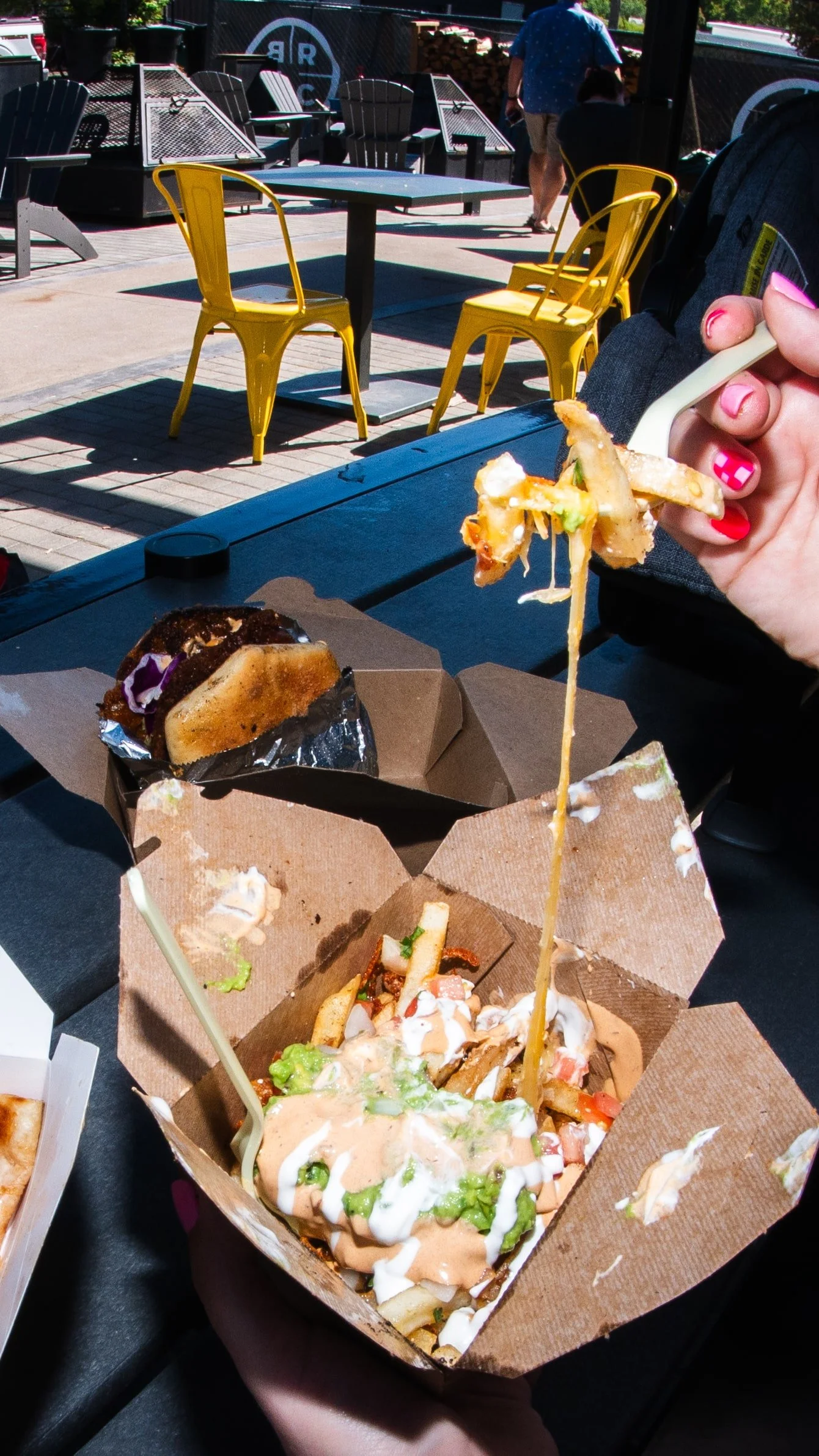 Outdoor setting with yellow chairs, black tables, and people in the background. Someone is holding a fork with French fries and toppings over a cardboard takeout container filled with fries, lettuce, tomatoes, and sauces, with another food item on th