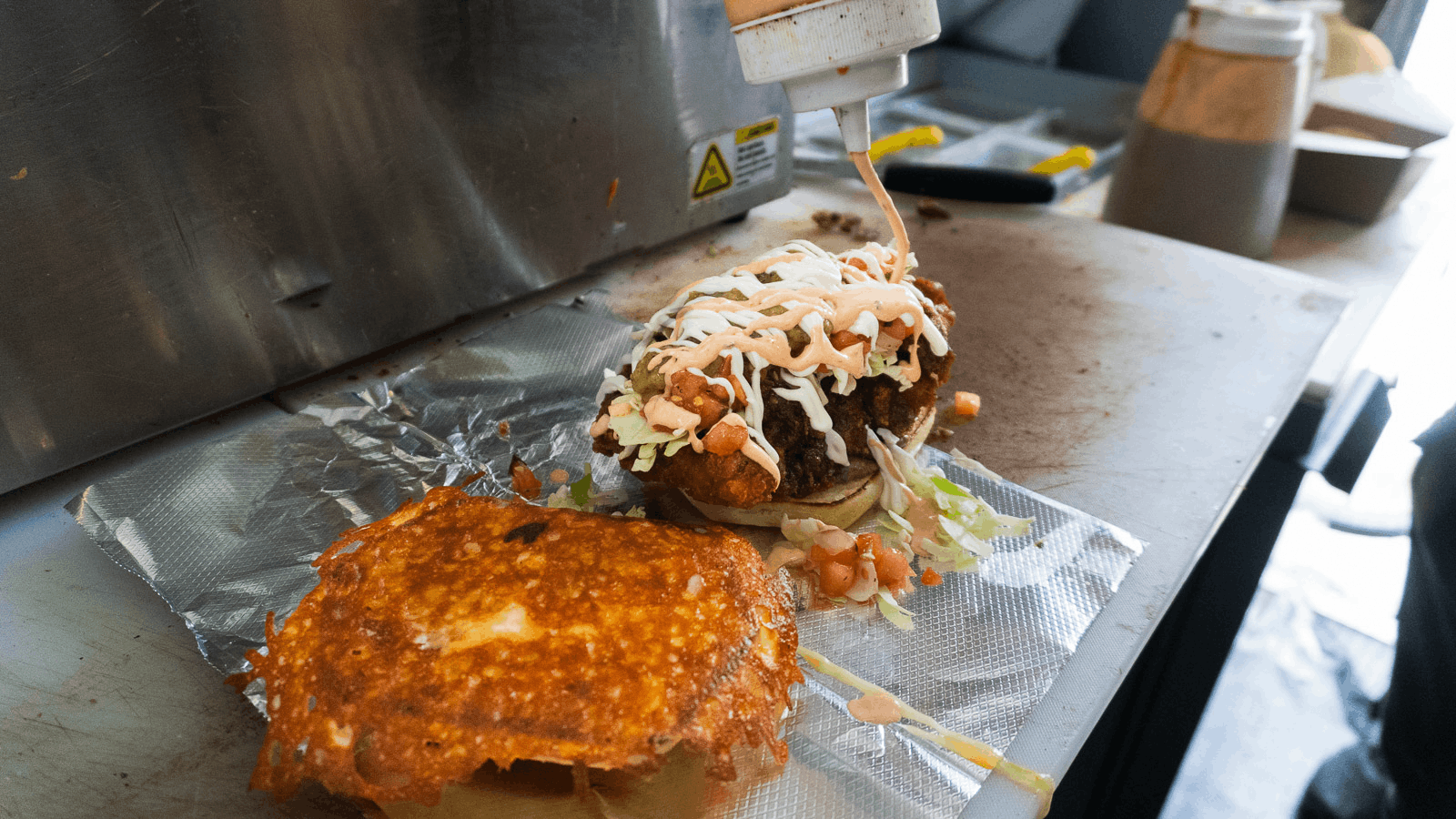 Fried chicken sandwich with toppings, and a piece of fried chicken on foil on a kitchen countertop.