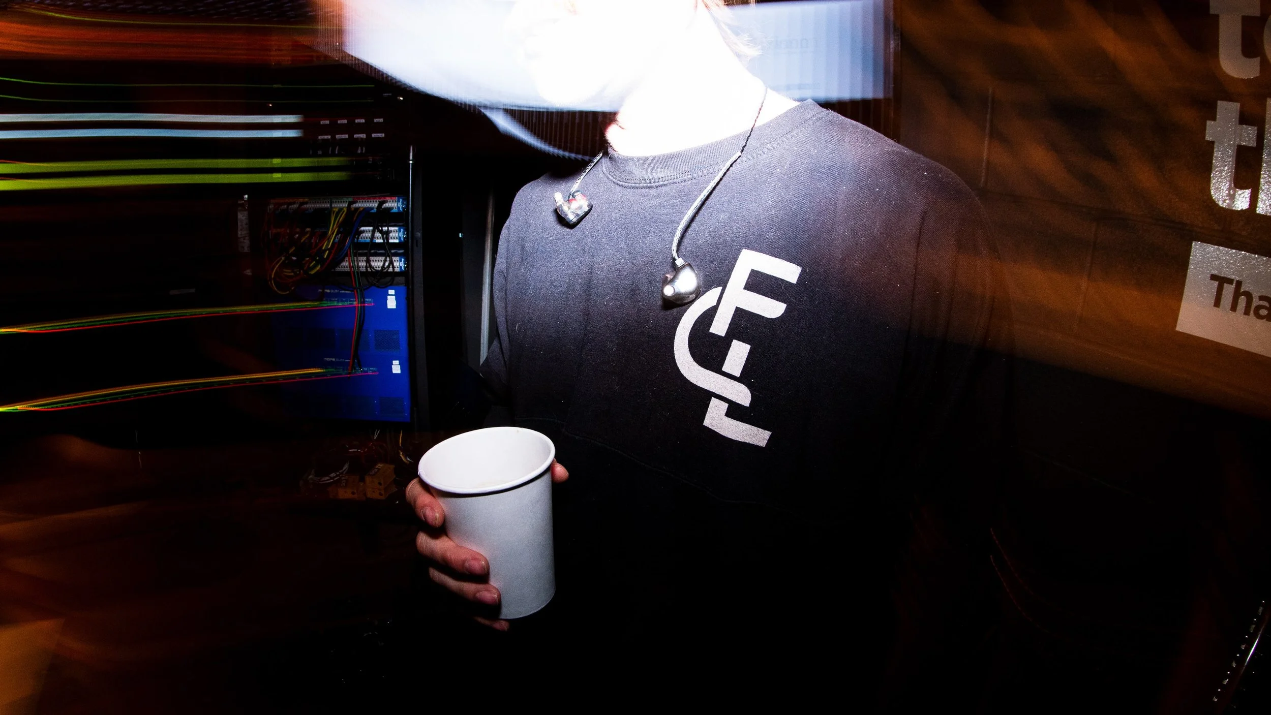 Person holding a white mug in front of a computer server with exposed wires, wearing a black shirt with white text and a necklace with black earbuds.