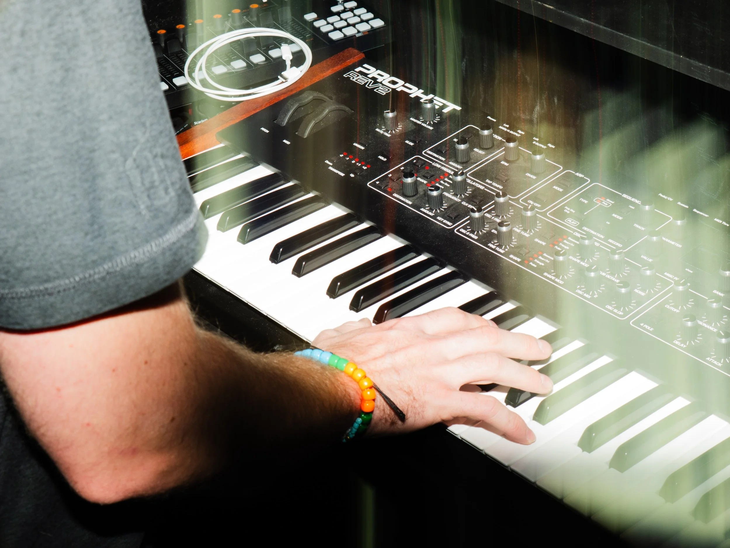 Close-up of a person's hand playing a vintage synthesizer keyboard, with various control knobs and a small screen visible, behind a glass panel, with a reflection of the person wearing a gray shirt and a rainbow bracelet.