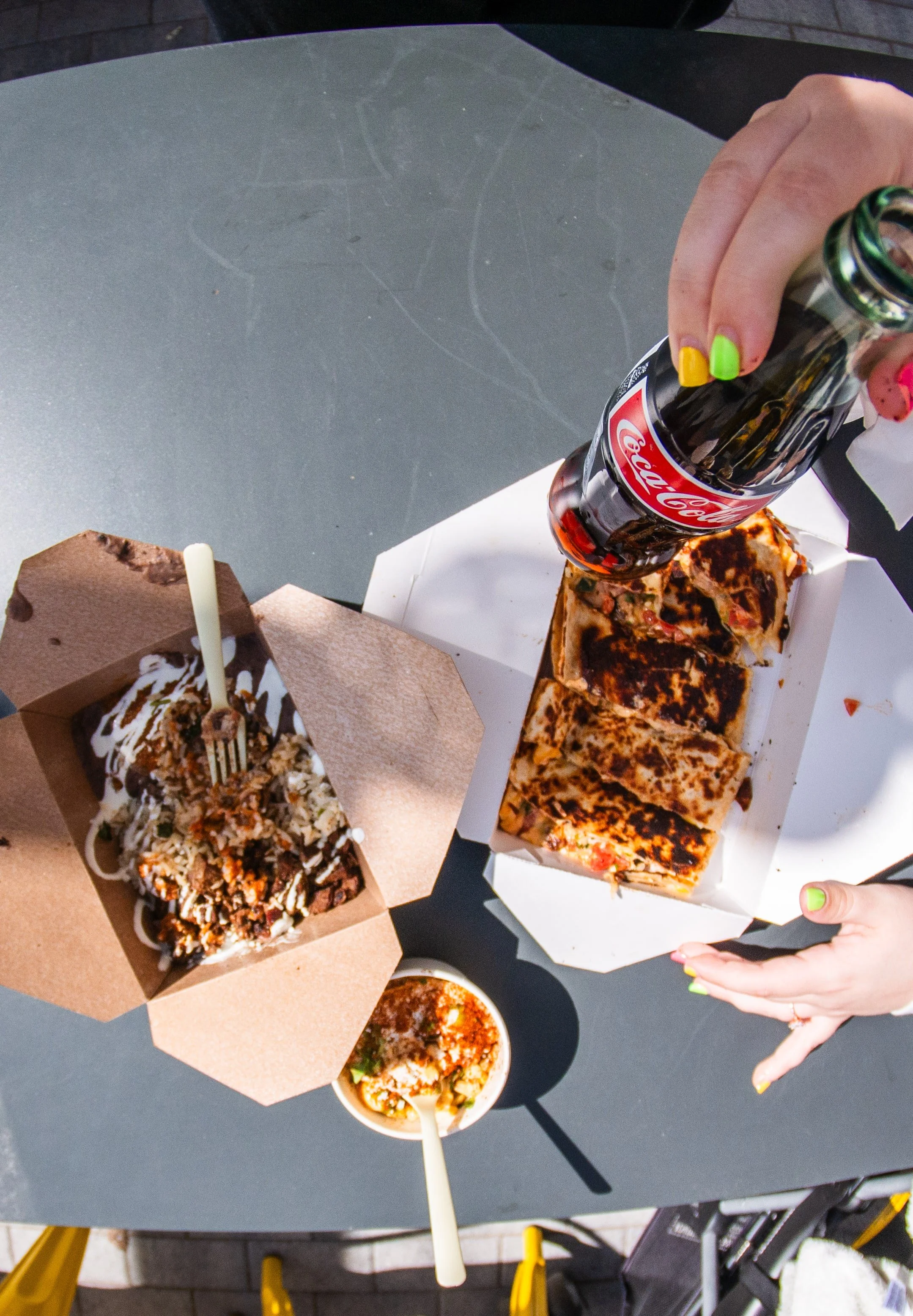Overhead view of a table with various fast food items, including a small cardboard box of loaded fries topped with cheese, sauce, and toppings, a white takeout container of slices of pizza with burnt crust, and a small cup of soup with a spoon inside