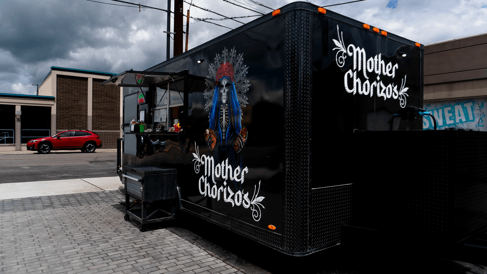 Black food truck with Mother Chorizo branding, featuring an artwork of a woman with blue hair and a skull face, parked on a city street.