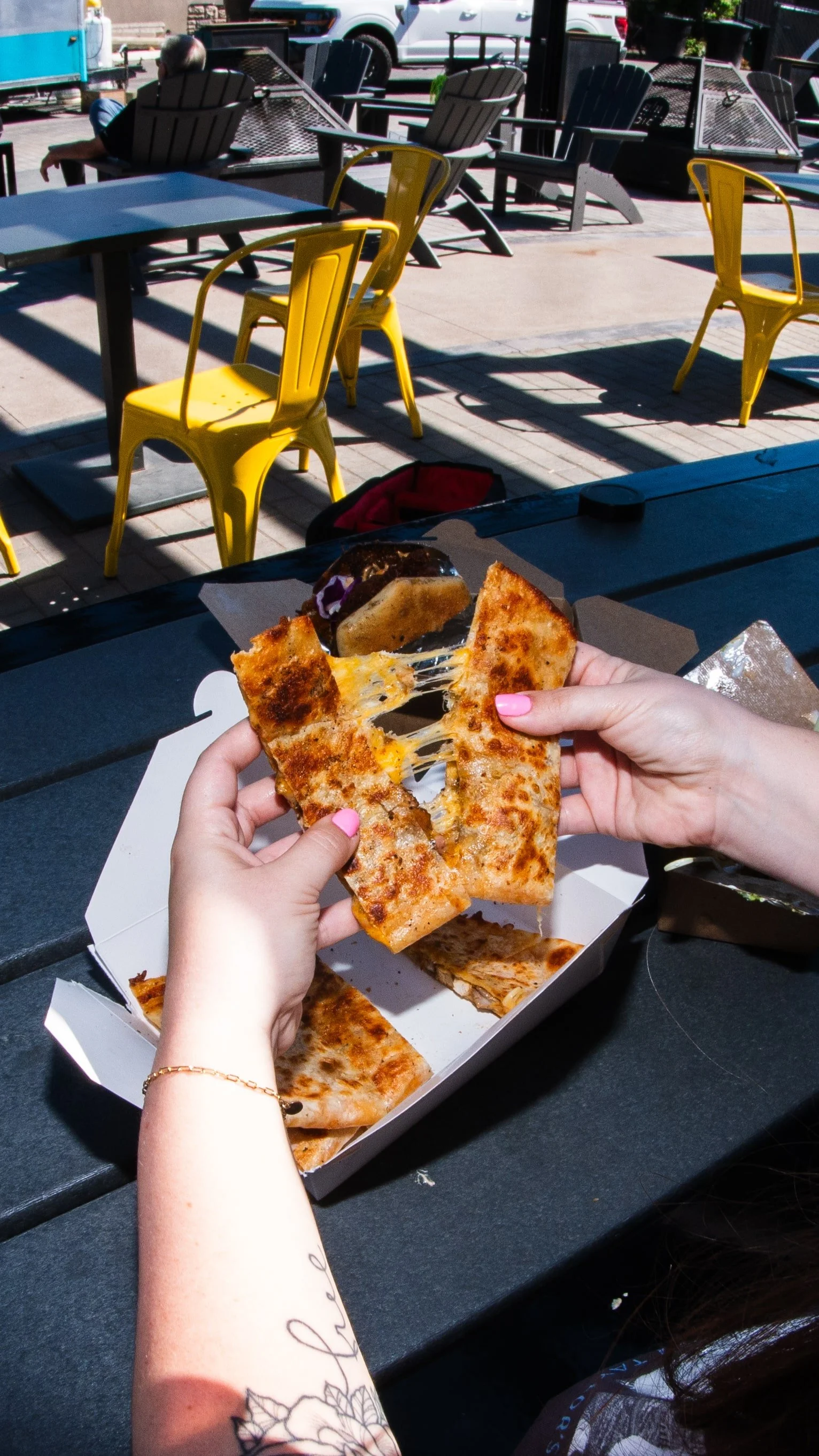 Person holding slices of pepperoni pizza, with melted cheese stretching between slices, at an outdoor cafe with yellow chairs and tables, and parked cars in the background.