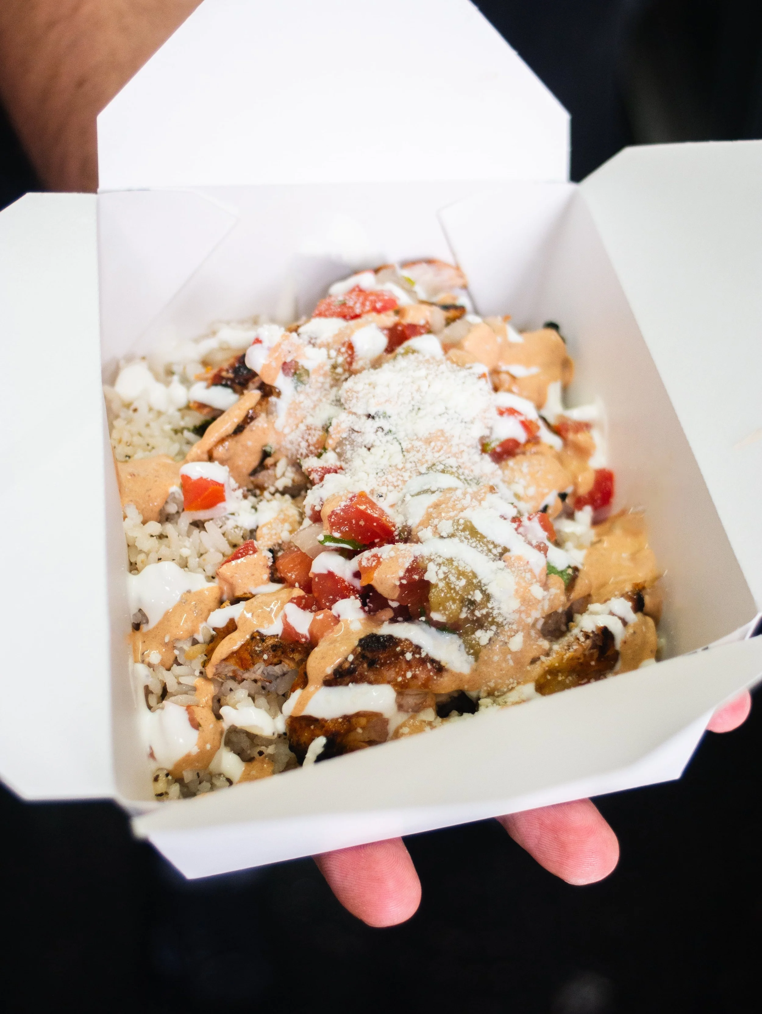 A white takeout container filled with rice topped with pieces of grilled chicken, diced tomatoes, white sauce, and grated cheese.