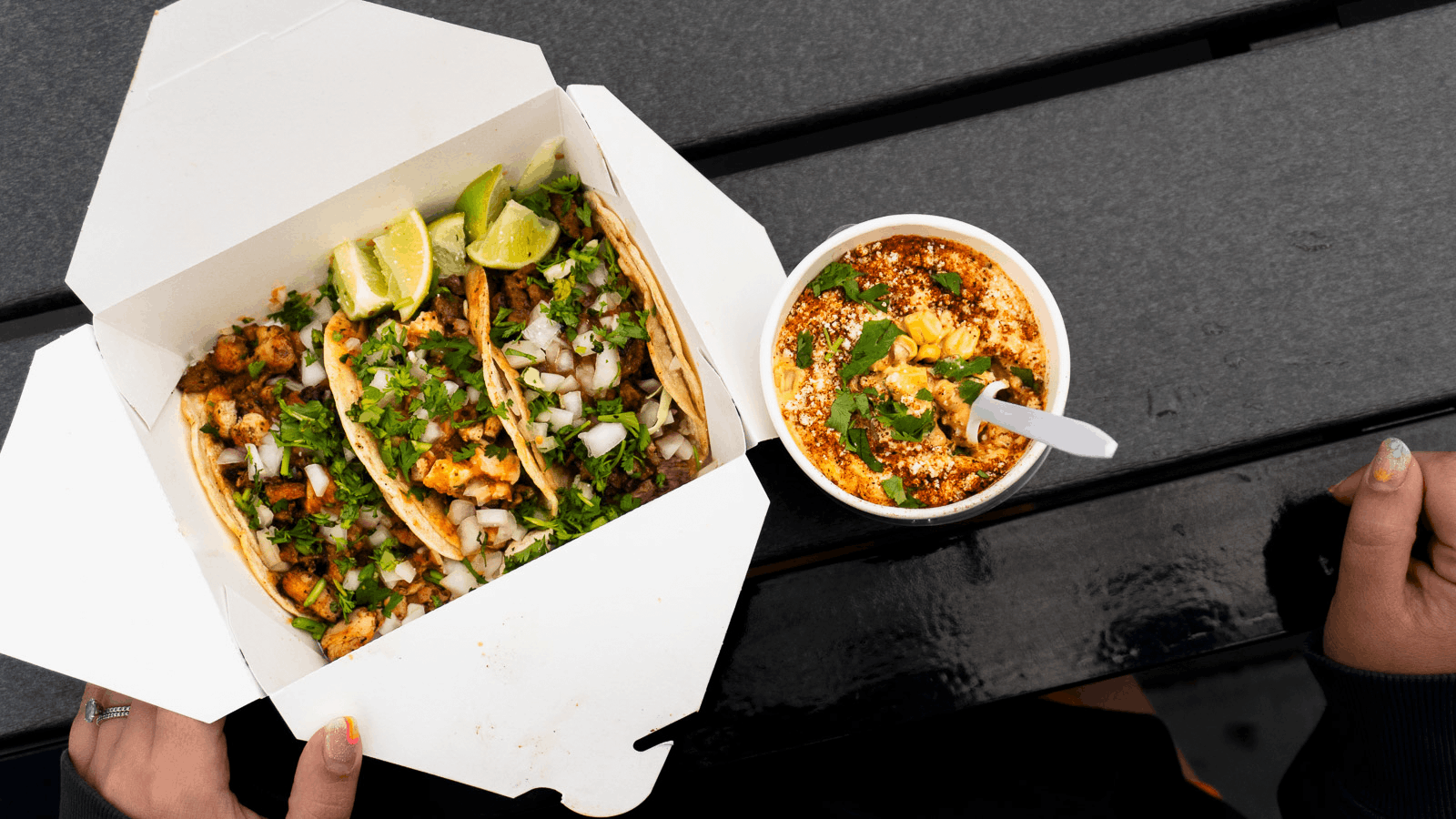 A top-down view of a hand holding a white food container with tacos filled with meat, chopped onions, and cilantro, topped with lime wedges, next to a cup of spicy soup garnished with cilantro and cheese on a black wooden table.