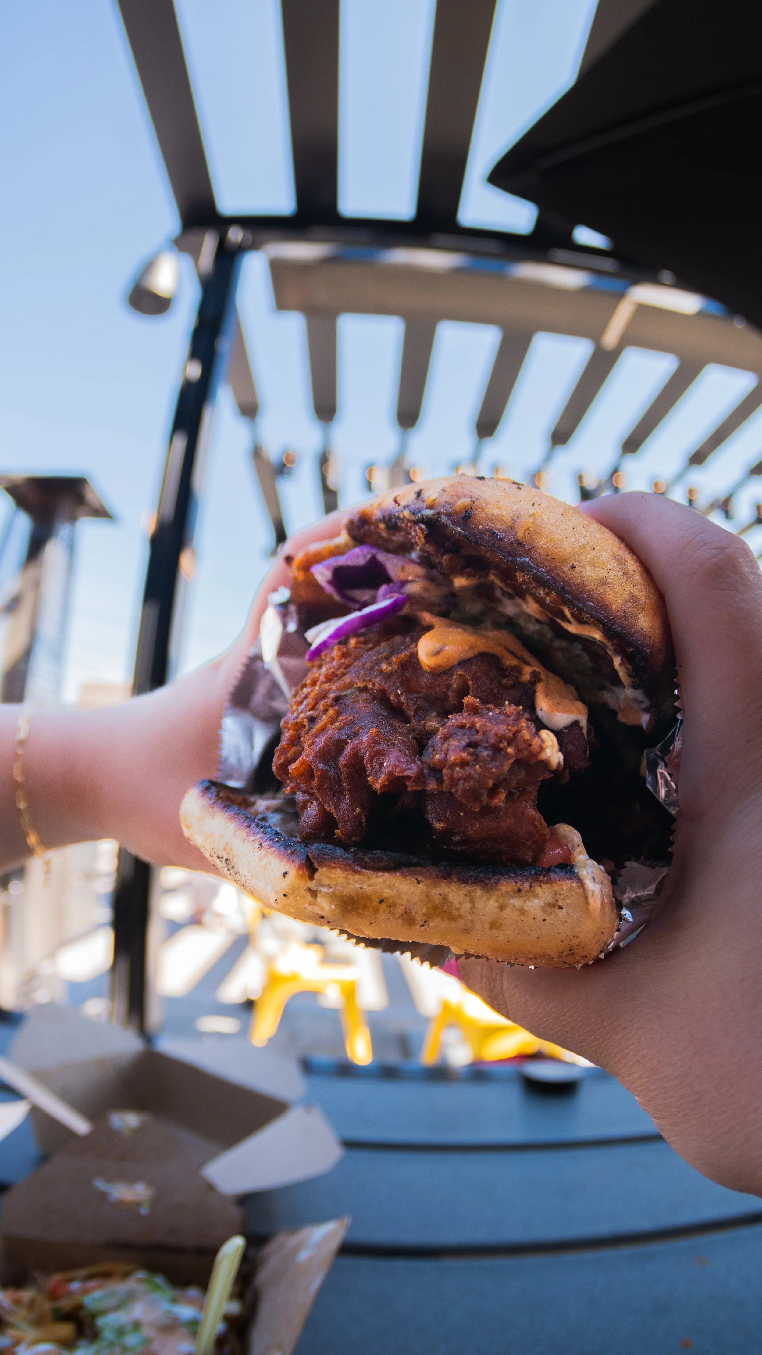 Person holding a fried chicken sandwich with purple cabbage and sauce at an outdoor setting with a striped canopy.