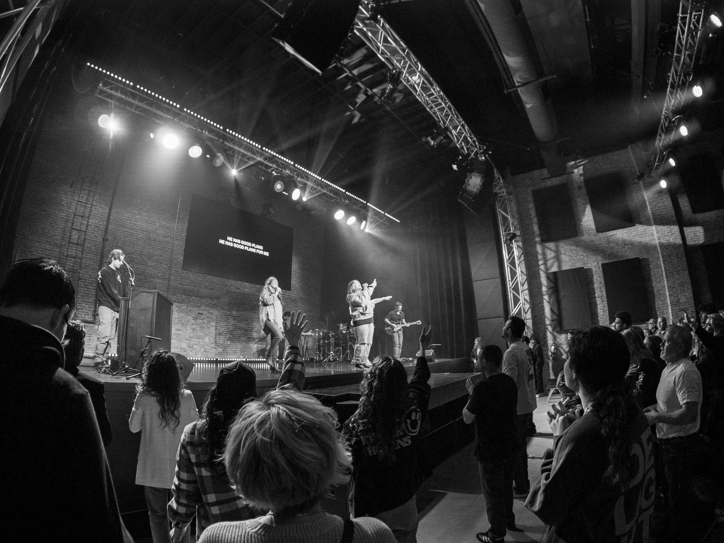 Black and white photo of a stage performance with a band and multiple people in the audience, some raising their hands, inside a venue with brick walls and stage lighting.