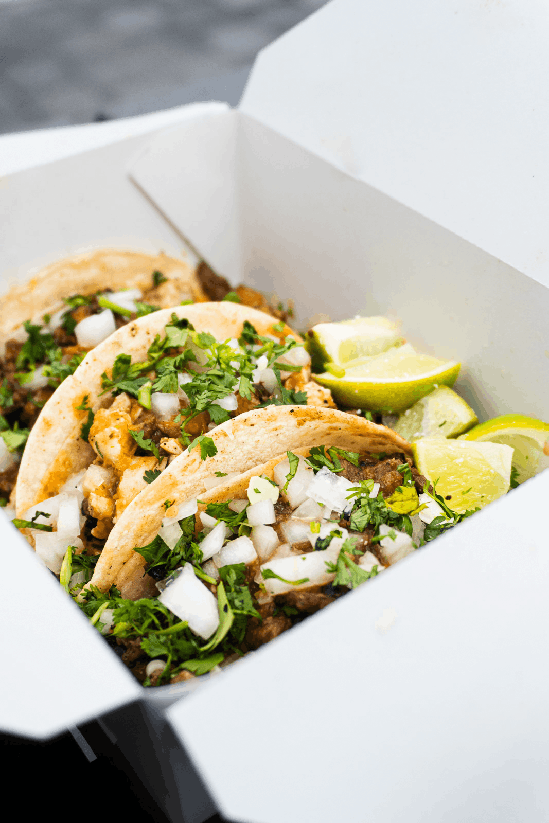 Three tacos with chopped onions and cilantro, served with lime wedges in a white takeout container.