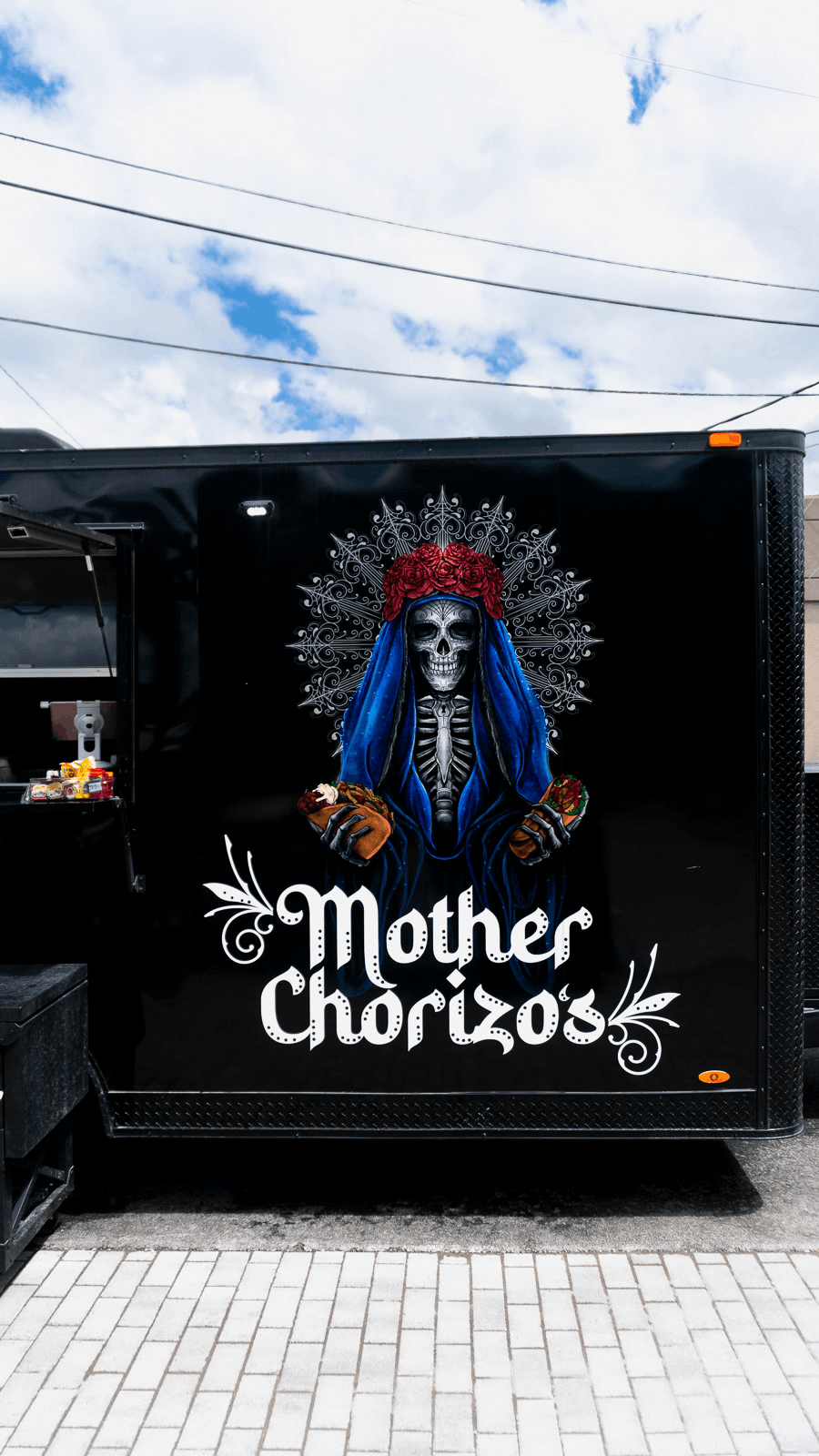 Artwork of a skeletal figure dressed as La Catrina with a red floral crown and blue cloak, holding tacos, with text 'Mother Chorizo' on a black background.