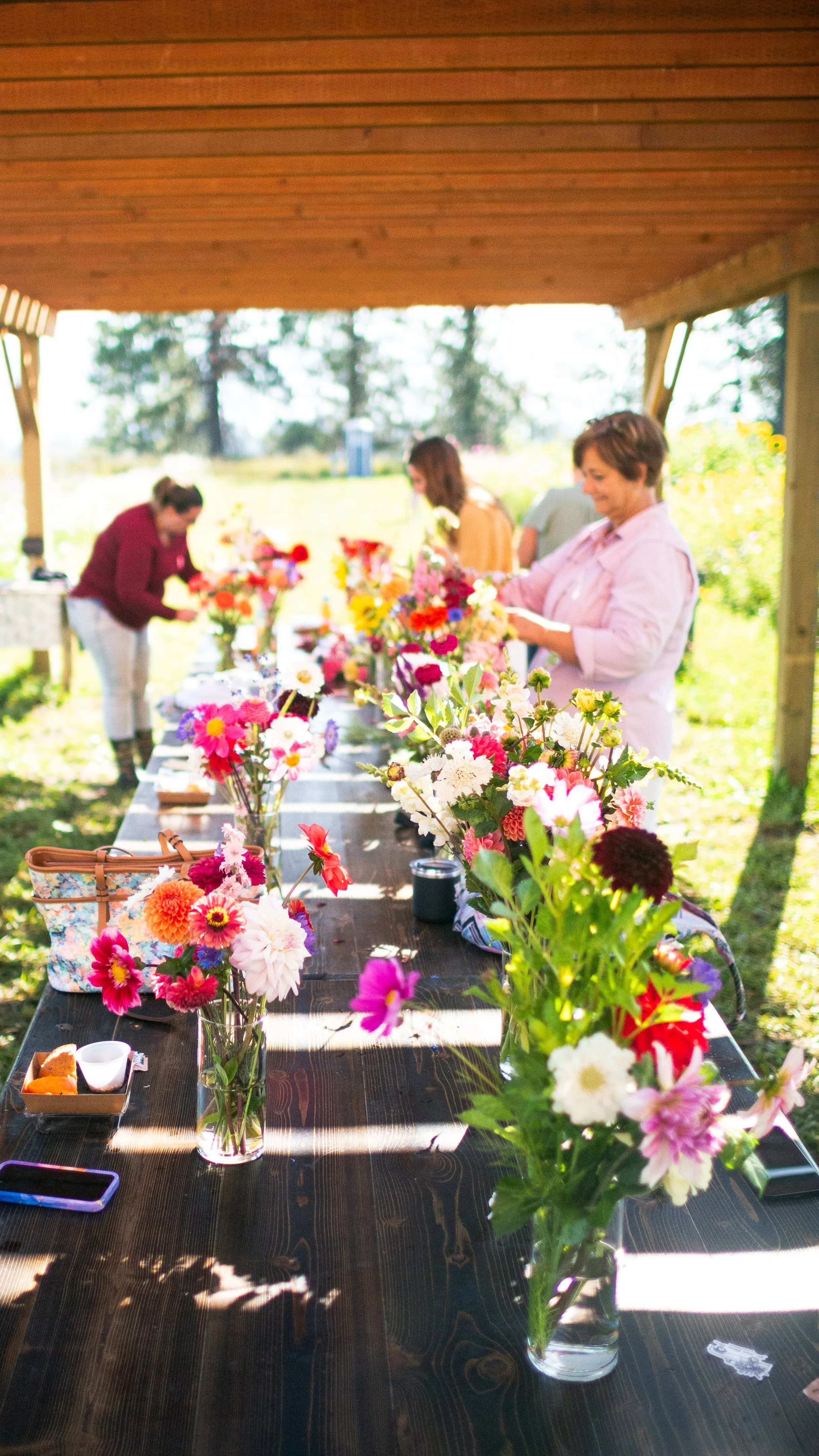 People arranging colorful flowers on a long outdoor table under a wooden roof.