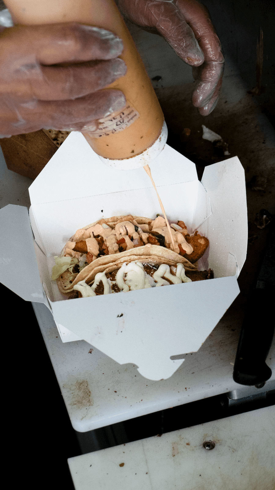 A person wearing gloves drizzles a sauce over a taco inside a white takeout box that contains multiple tacos, with visible toppings like lettuce, cheese, and meat.