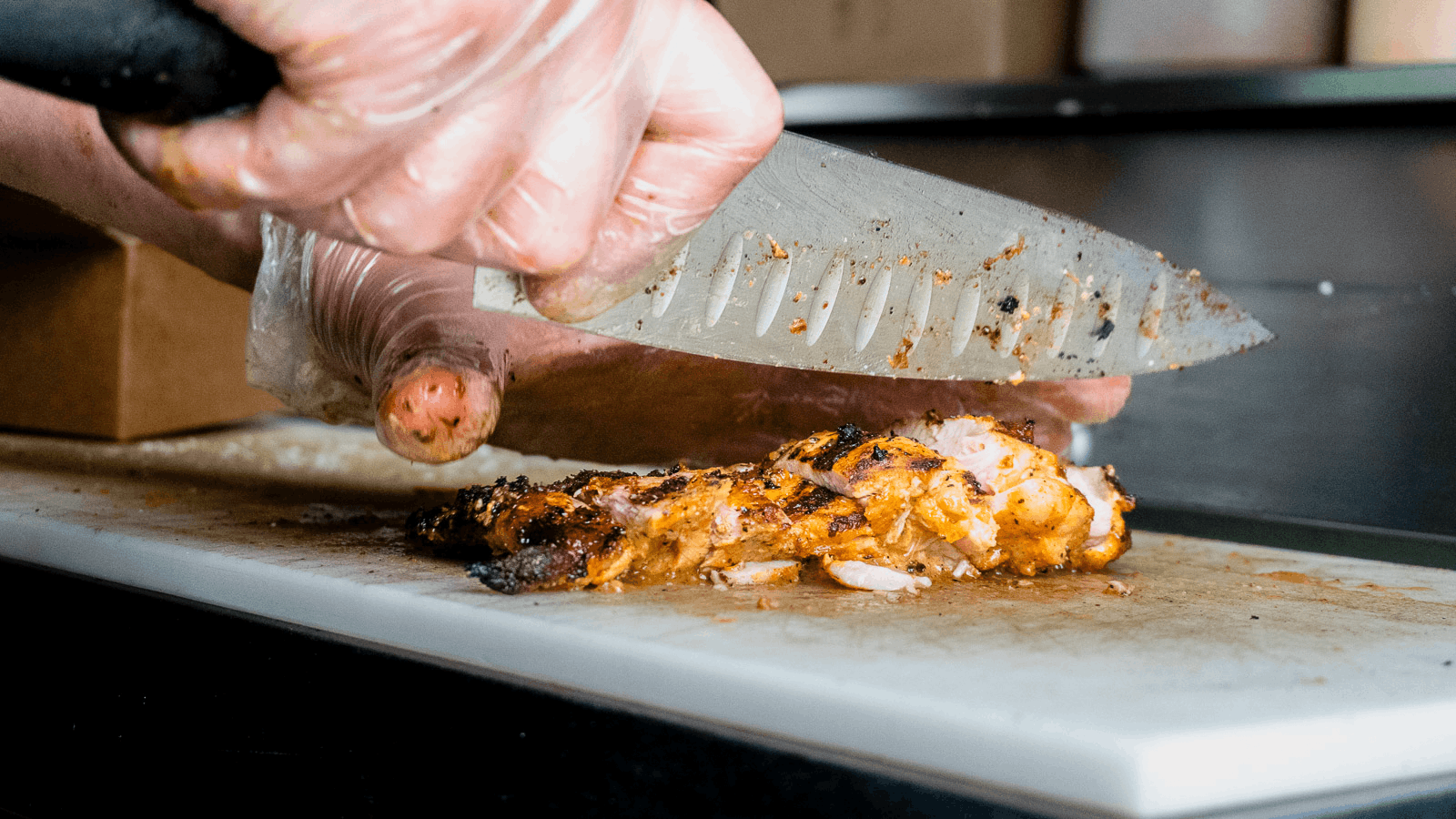A person wearing gloves cuts grilled chicken on a cutting board with a large knife.