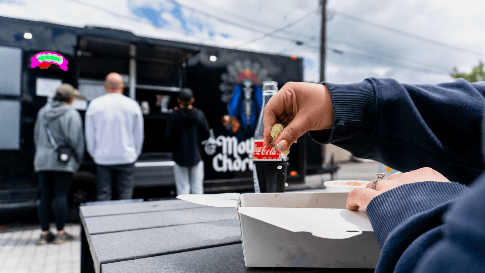 Person opening a food container with a small drink at a food truck, with three people waiting in line in the background.