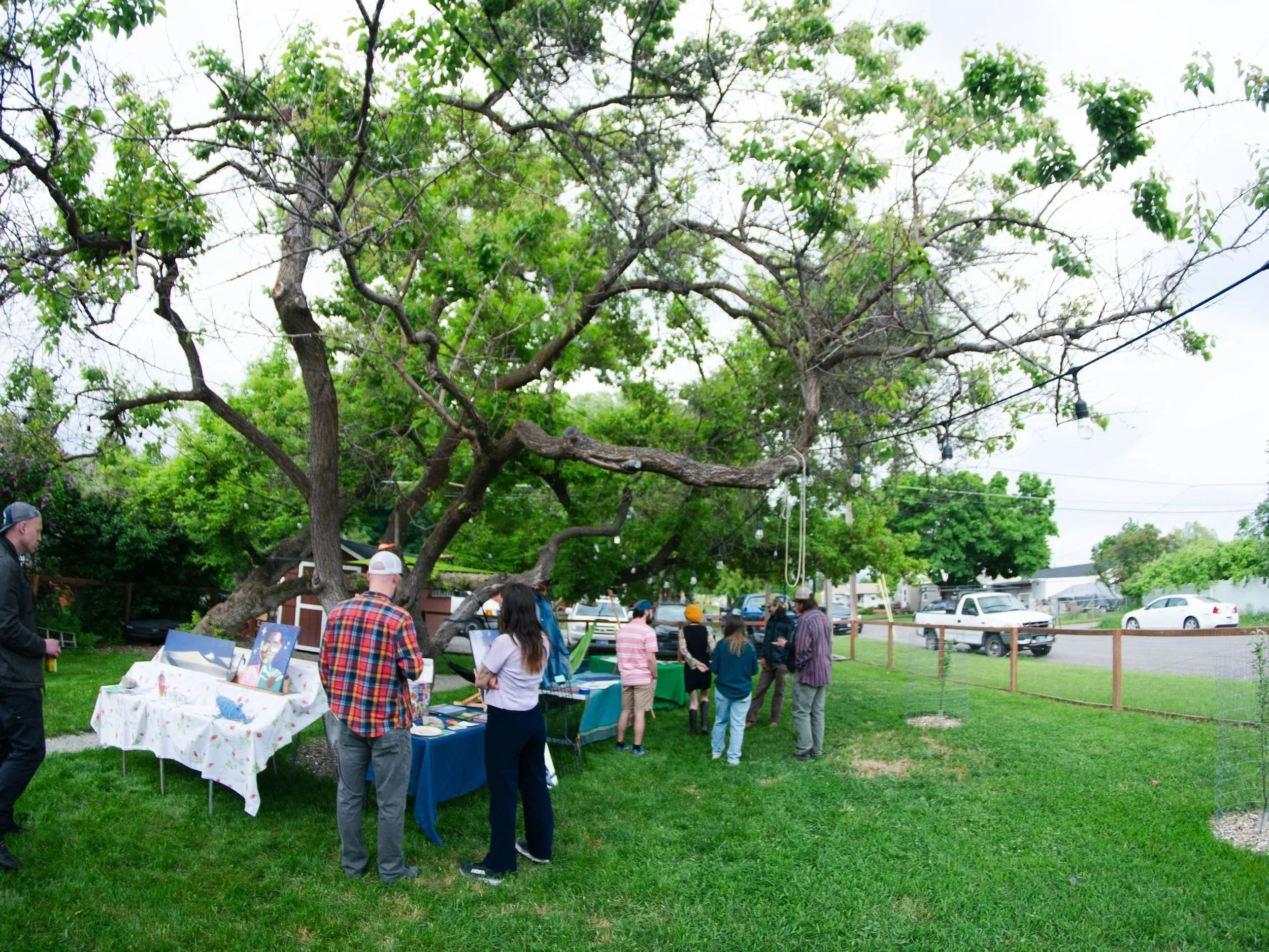 People browsing art and craft tables under a large tree in an outdoor yard sale or market.