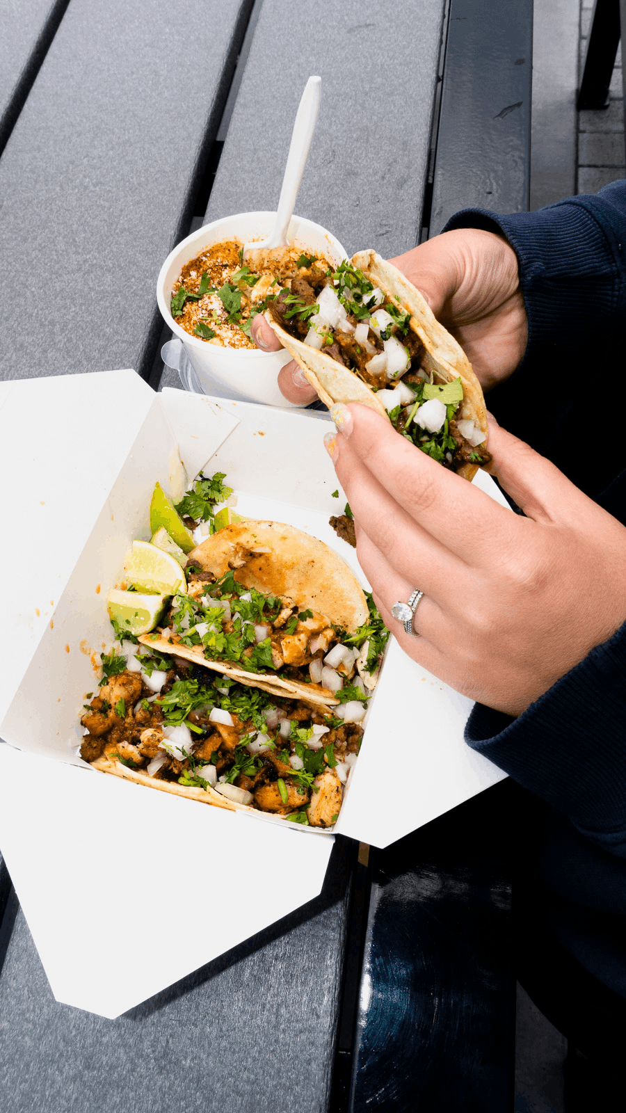 A person holding a street-style taco filled with seasoned meat, chopped onions, and cilantro, with lime wedges and more tacos in a takeout box on a gray table.
