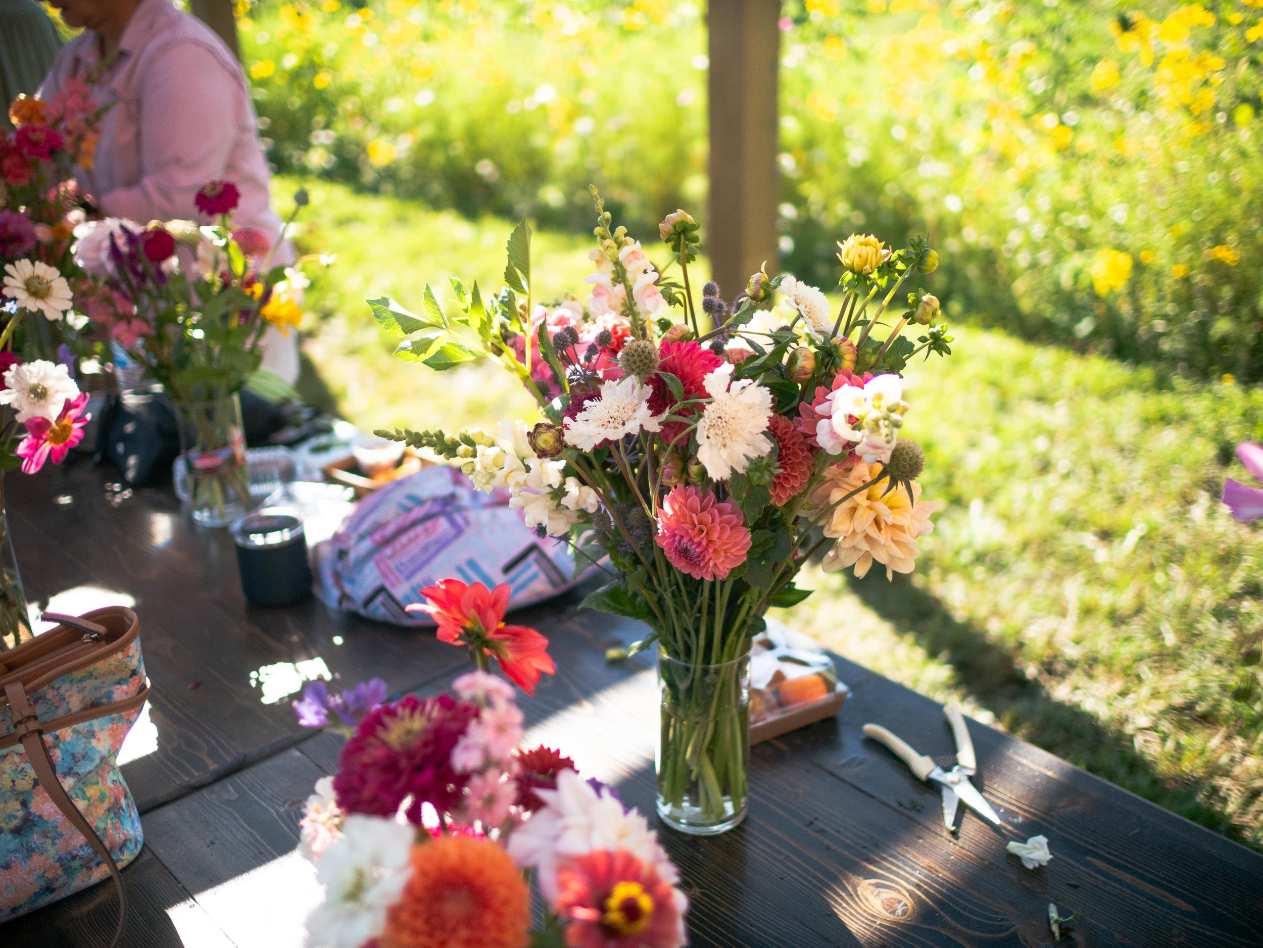 A large bouquet of flowers in a glass vase on a wooden table outdoors with a sunny garden in the background.
