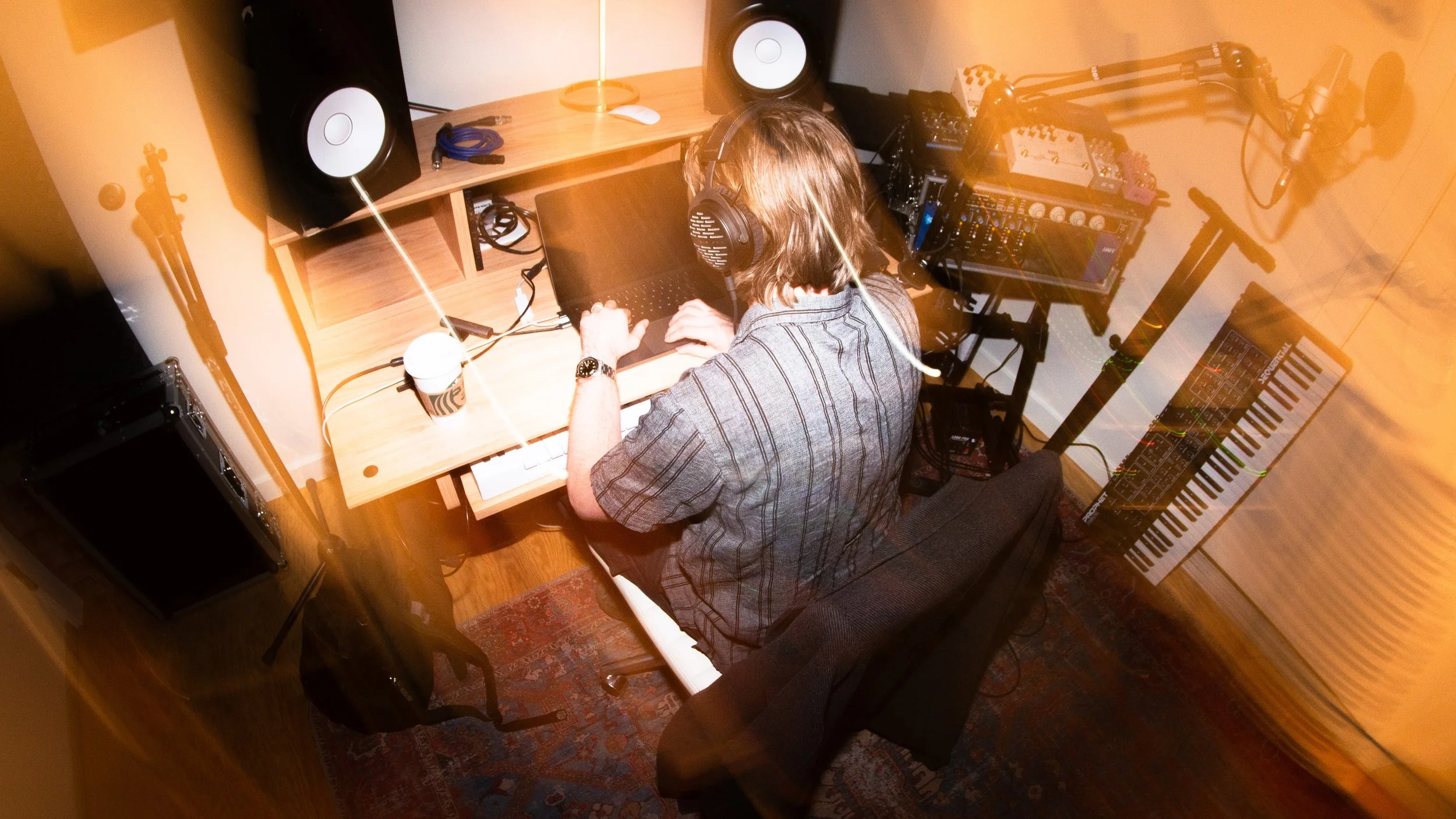 A person with shoulder-length hair is sitting at a desk, wearing headphones, and working on a laptop surrounded by music equipment including studio speakers, a keyboard, and a mixer in a home studio.