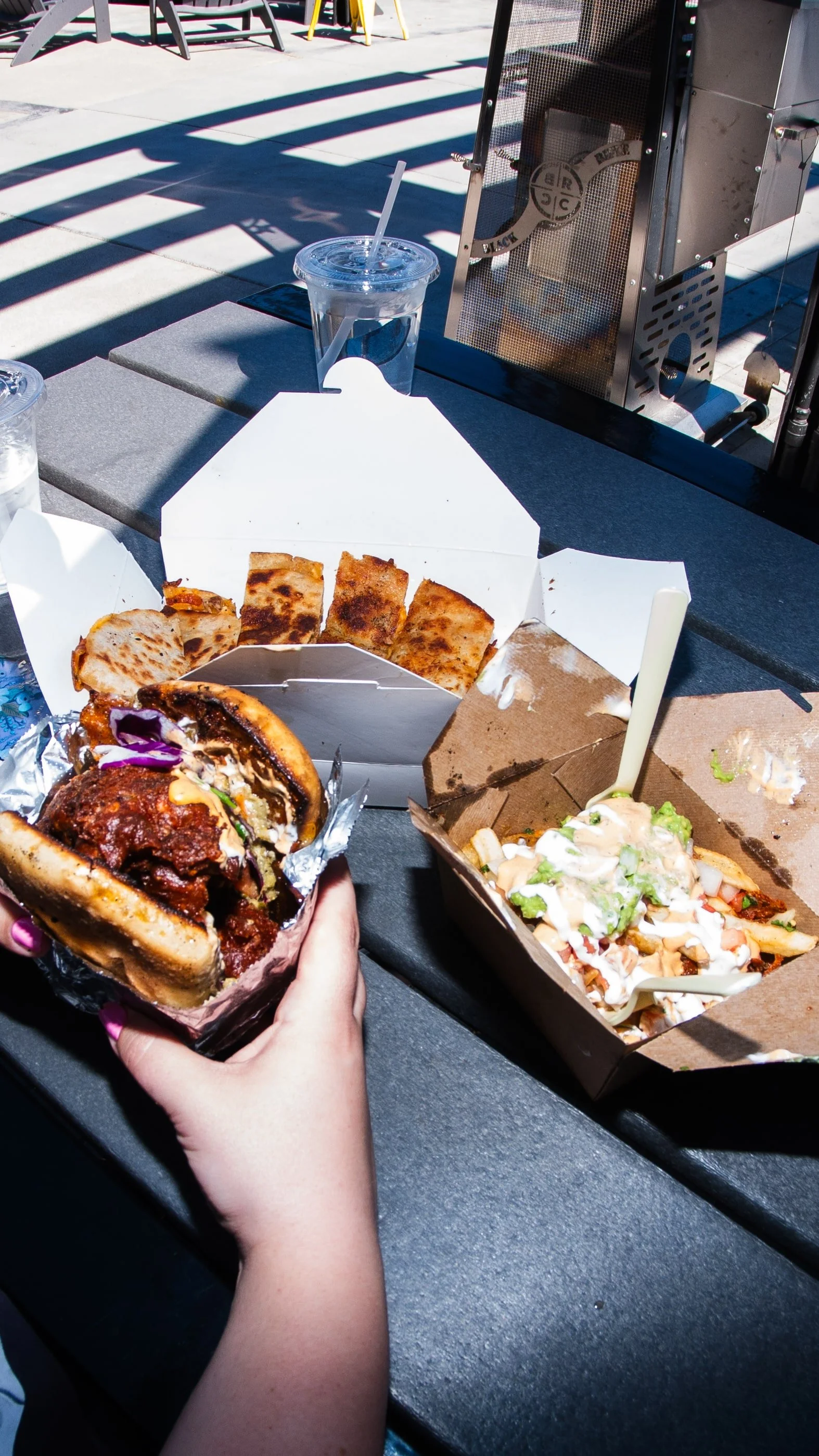 A close-up of a hand holding a burger with fried chicken, lettuce, and sauce in a paper wrapper. On a black outdoor table, there are also pizza slices in a white box, seasoned fries in a brown box with dressing and lettuce, two cups of iced water, an