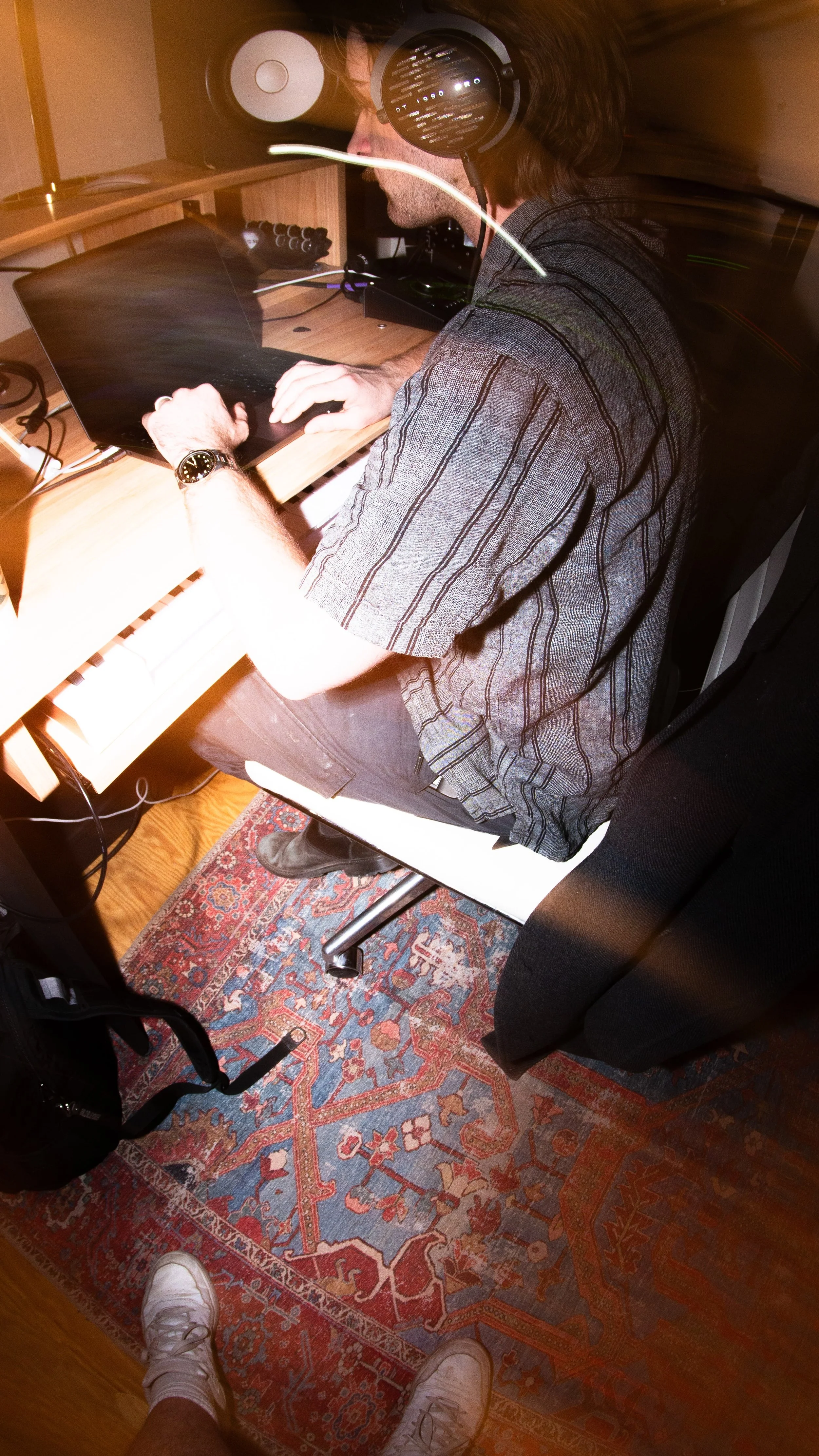 A man with long hair and a striped shirt wearing headphones working at a digital music production setup, sitting on a white chair in a room with a wooden floor and a patterned rug.