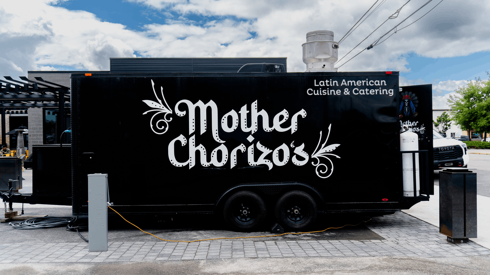 Black food truck with large white lettering reading 'Mother Chorizo' and smaller text 'Latin American Cuisine & Catering'; parked outdoors under a partly cloudy sky.