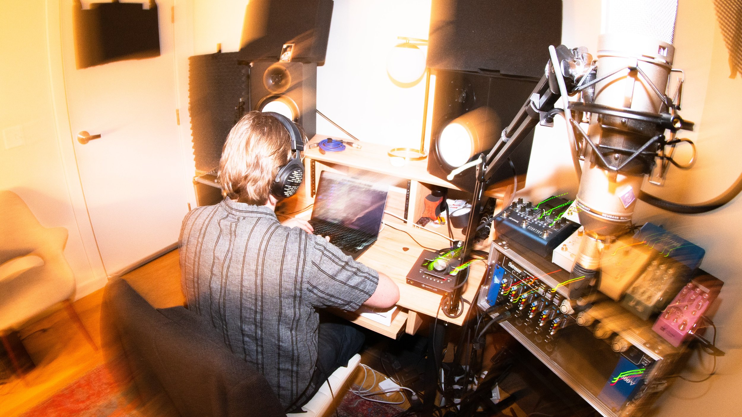 A person with shoulder-length hair wearing headphones working on a laptop in a home studio with musical equipment and monitors.