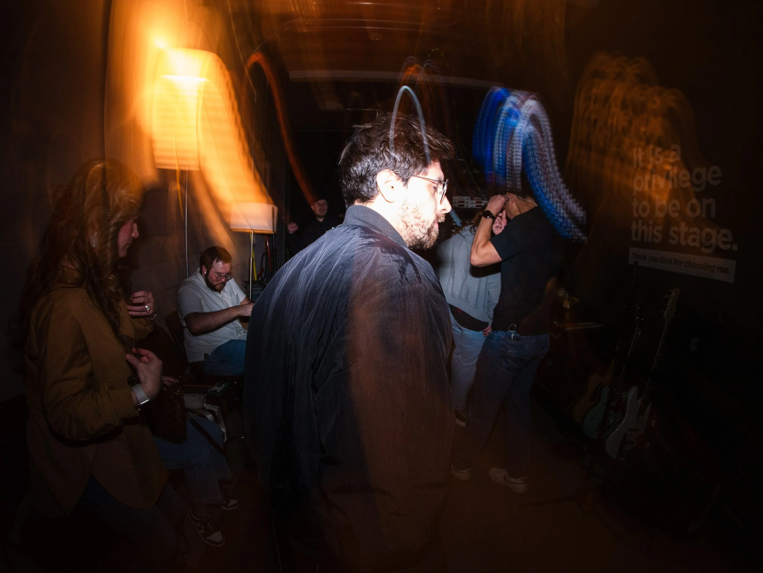 People praying in a dimly lit room with multicolored light streaks, guitar on stand, and a sign that says 'Thank you God for choosing me.'