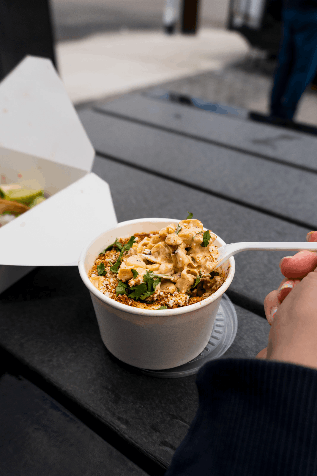 A person holding a spoonful of creamy mac and cheese in a white paper cup outdoors on a black table.