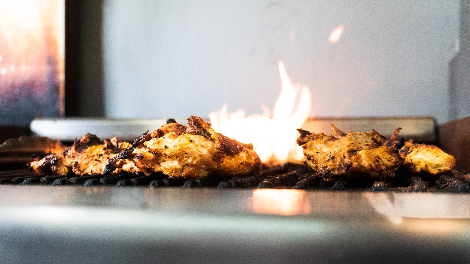 Chicken pieces cooking on a grill with flames in the background, inside a kitchen oven.
